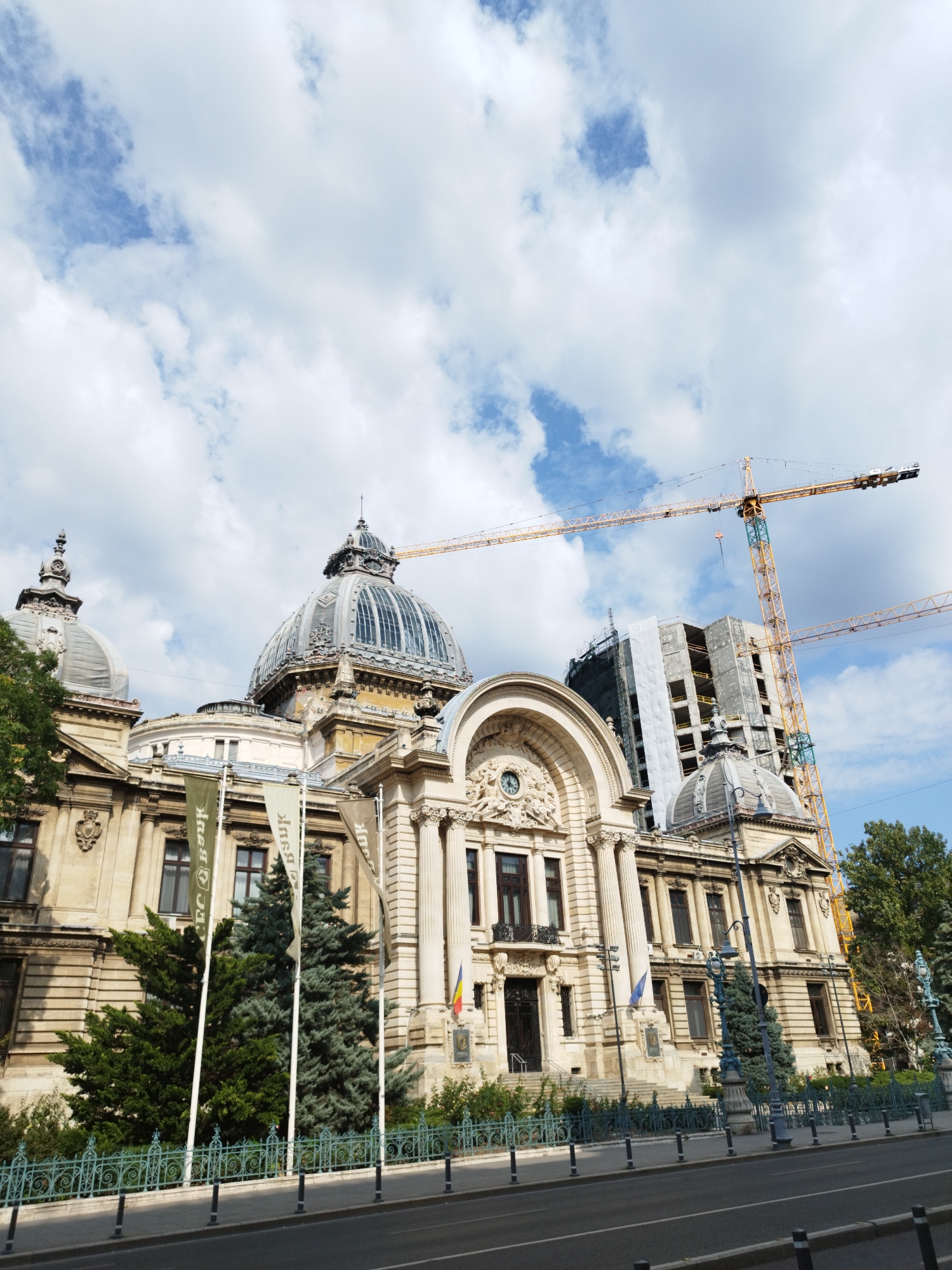 This is a low-angle, wide shot of a grand, ornate building with domes and columns, likely a historical landmark in Bucharest, Romania. The building appears to be constructed of light-colored stone and features intricate architectural details. In the foreground, there is a street with a metal fence bordering a sidewalk. Several tall, green evergreen trees are planted behind the fence, and three tall flags are erected in front of the building, partially obscuring it. Two large yellow construction cranes are visible in the sky behind the building, with a partially constructed, modern building also under construction. The sky is partially cloudy with patches of blue. The overall impression is a juxtaposition of old-world architecture and modern development.
