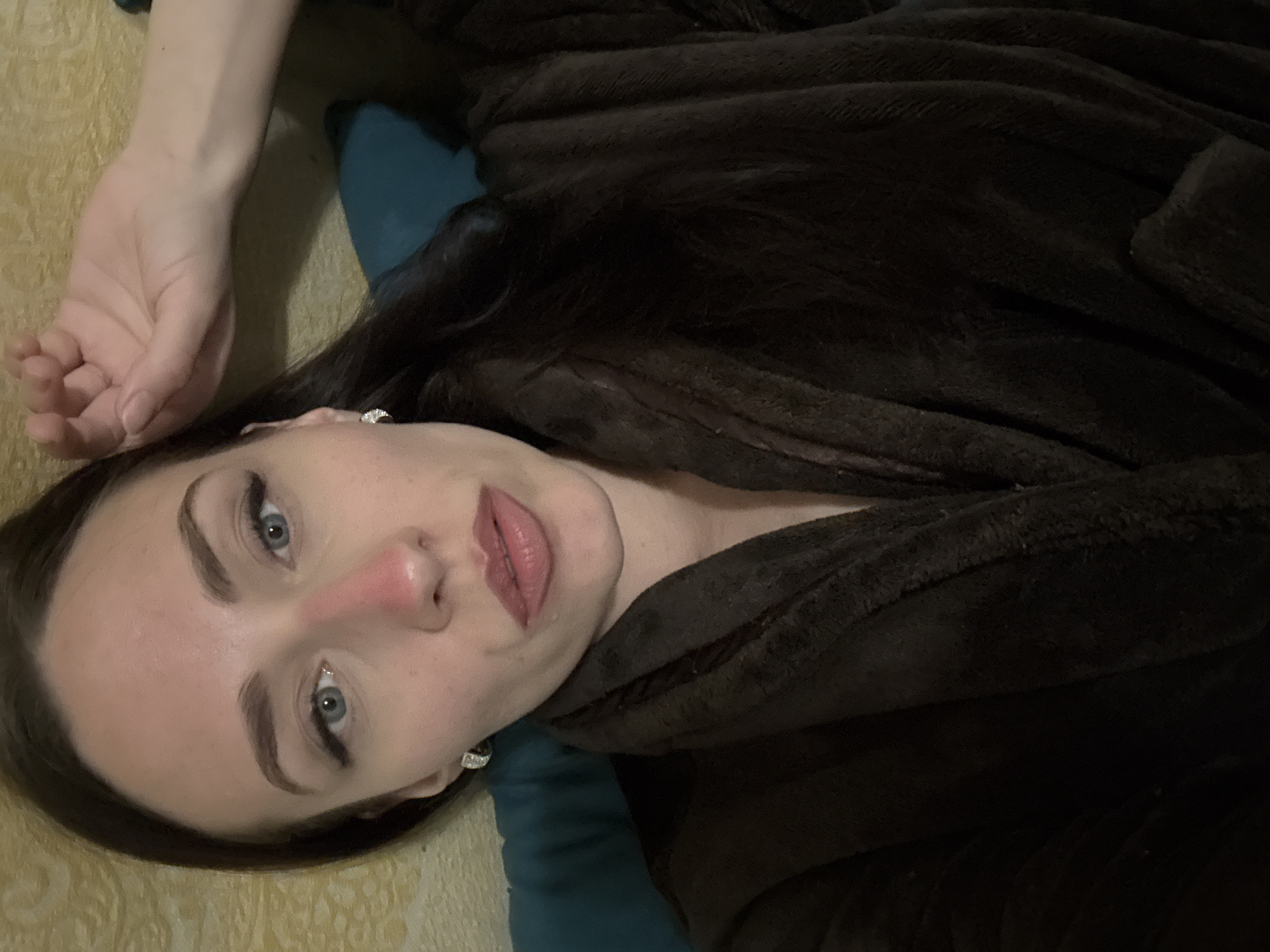 A woman lies on her back wearing a dark brown, possibly velvet, robe. She is resting on a blue pillow atop a patterned surface. Her face is visible, with fair skin, light blue eyes, and dark hair. She is wearing earrings. Her left arm is extended above her head. The location cannot be determined from the image.