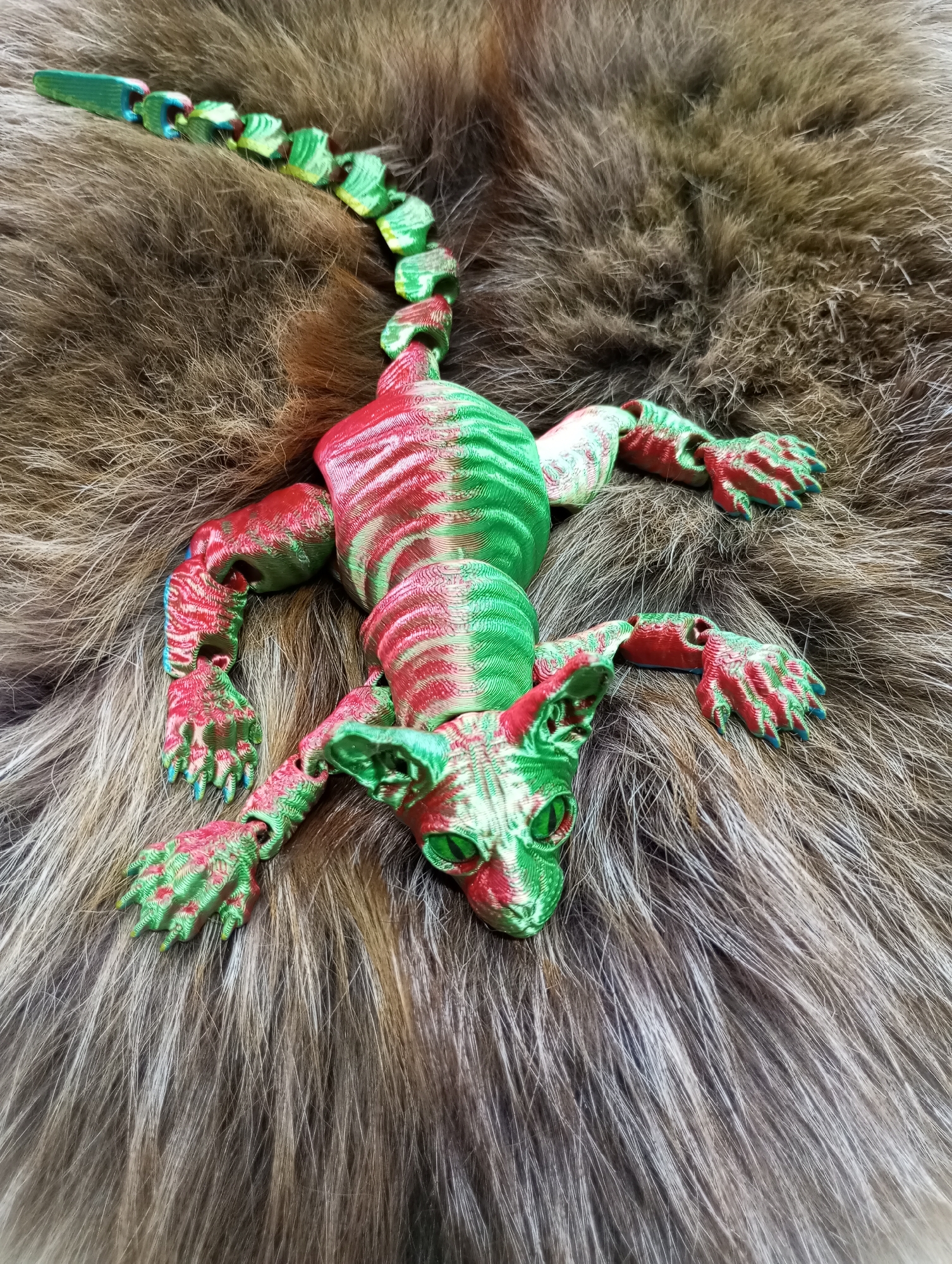A 3D-printed, flexible toy depicting a creature resembling a cat-lizard hybrid is resting on a brown, furry surface. The toy is multicolored, primarily green and red, with articulated limbs and a segmented tail. The furry surface appears to be a type of animal fur or faux fur fabric. No elements in the image suggest a location in Pervomaysky, Russia. The image focuses on the texture of the toy and the fur.