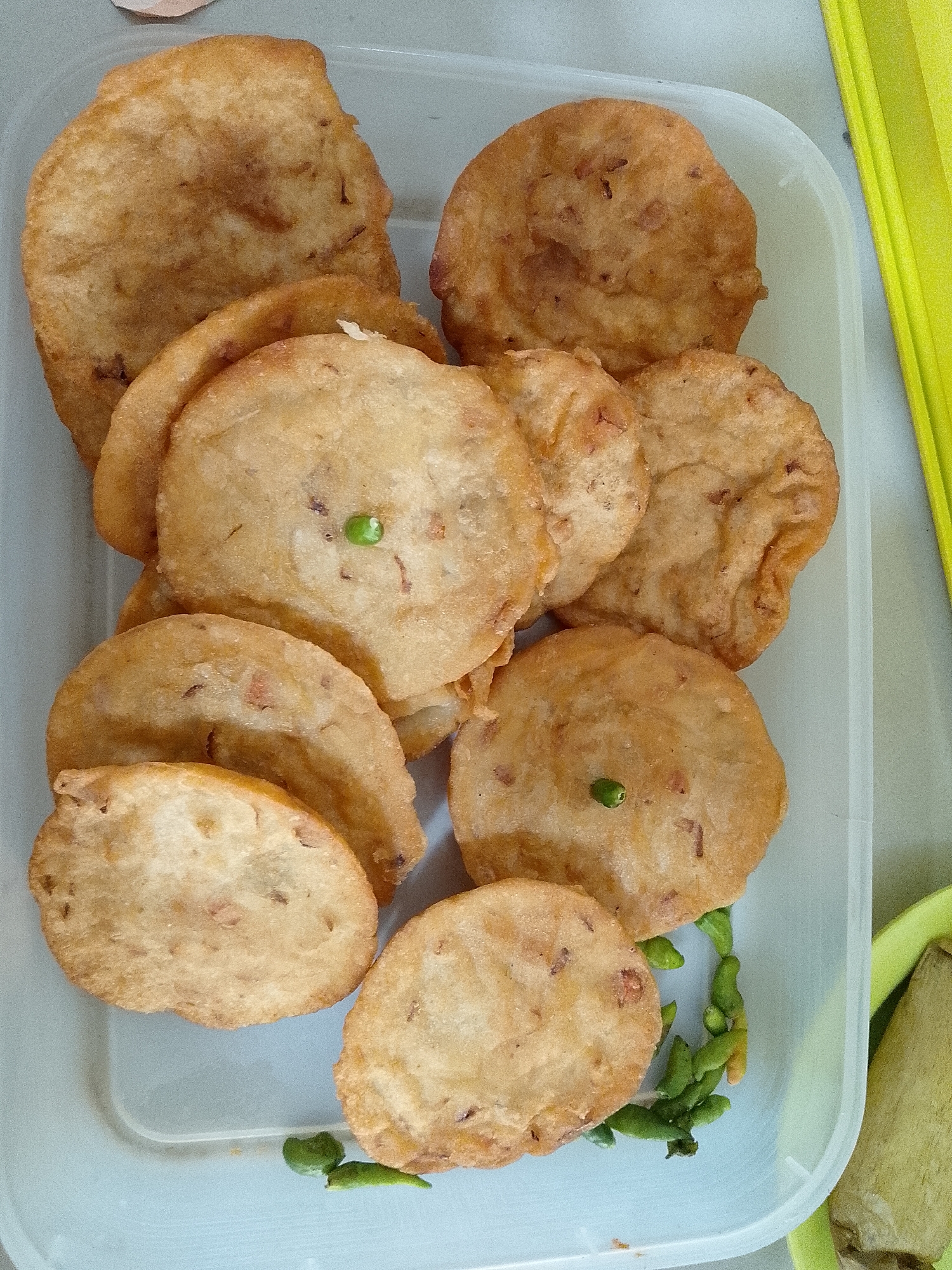 In Lhokseumawe, Indonesia, a close-up overhead shot captures a clear plastic container filled with an assortment of fried fritters. The fritters are predominantly round and flat with irregular edges, varying in shades of golden brown, indicating they have been deep-fried. Some fritters exhibit visible flecks of what appear to be small pieces of vegetables or protein within their batter. One fritter in the center has a single small, green chili pepper resting on its surface. Scattered around the fritters, particularly towards the right side of the container, are several small, whole green chili peppers. A bright yellow object, possibly a placemat or cloth, is partially visible on the right edge of the frame. The background is a muted gray surface, suggesting an indoor setting.
