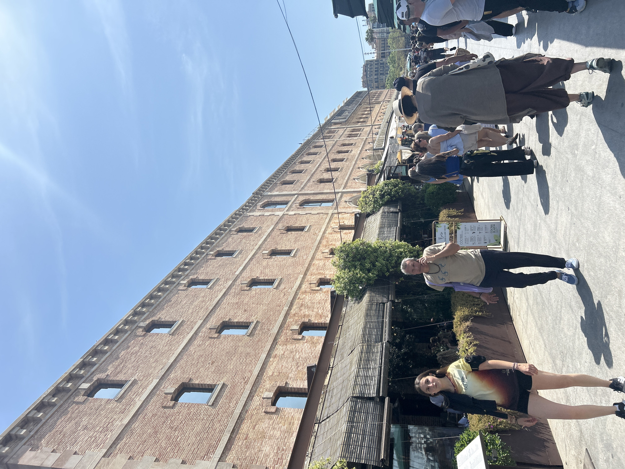 A sideways view shows a multi-story brick building on a clear day. Several people are seen walking along the sidewalk in front of the building. A tree and some foliage are partially visible in front of the building, along with awnings and umbrellas. The sky is clear and blue. Based on the architecture of the building, the image depicts the Port Vell area in Barcelona, Spain, which formerly served as the city's harbor.