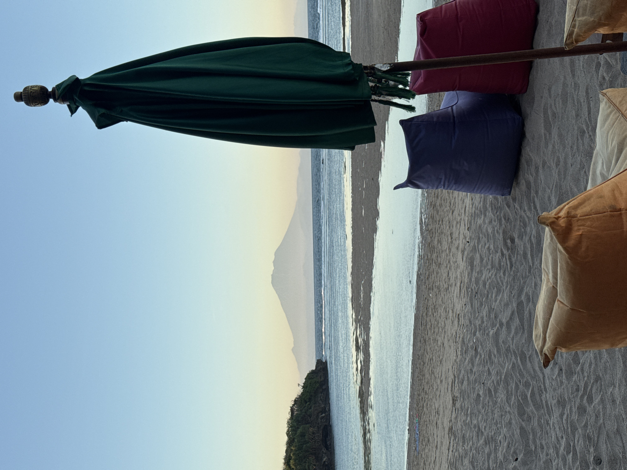 The image shows a beach scene in Kuta Selatan, Indonesia. A closed green umbrella hangs to the left of the frame. Several colored beanbag chairs (red, blue, tan) are arranged on the sand. In the background, the ocean meets the shoreline, and a mountain is visible in the distance under a clear sky. There are no people in the frame. Vegetation is present on the left side of the frame, between the beach and the ocean.