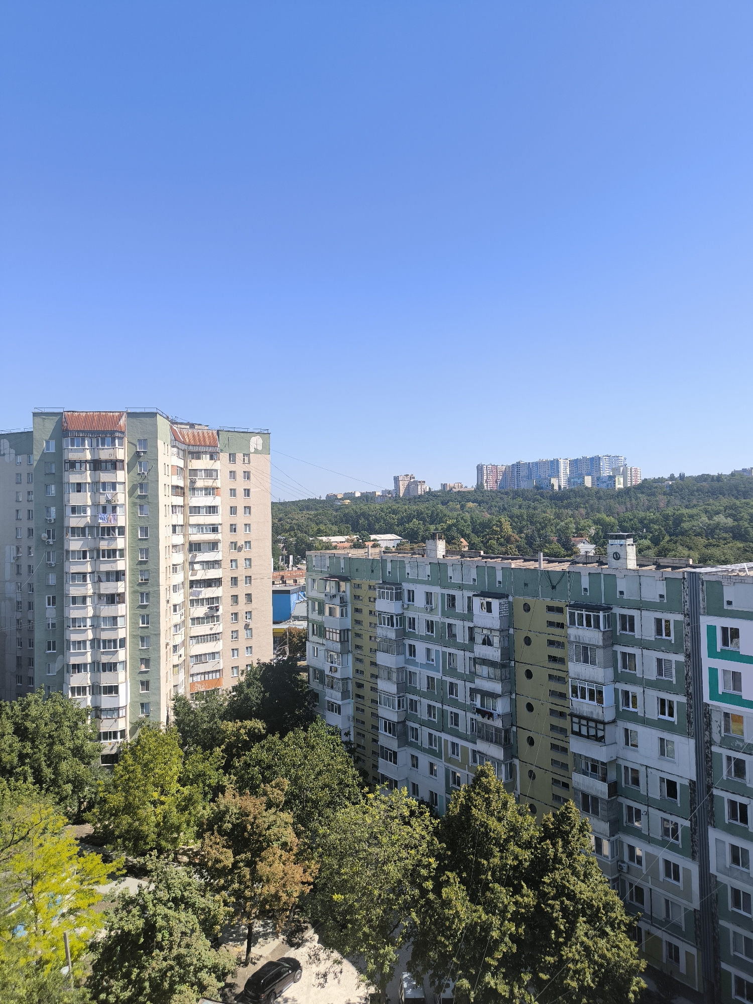 This is a photograph taken from a high vantage point overlooking a residential area in Chișinău, Moldova. The image features several multi-story apartment buildings, characteristic of Soviet-era architecture, with balconies and numerous windows. The buildings are painted in muted colors, primarily light gray, green, and beige, with some showing signs of wear and tear, such as rust-colored roofs on one of the buildings.
In the foreground, lush green trees partially obscure the lower floors of the apartment buildings, suggesting a neighborhood with established greenery. A black car is visible parked on a street below, surrounded by more trees.
In the background, a dense forest stretches across rolling hills. Beyond the trees, more apartment complexes and taller buildings can be seen on the horizon, indicating the urban sprawl of the city. The sky is a clear, bright blue, suggesting a sunny day. The overall scene depicts a typical urban landscape in Chișinău, with a blend of residential structures, natural elements, and distant city development.