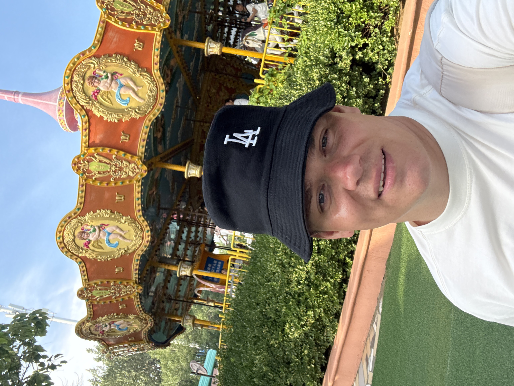 A man wearing a white t-shirt and a black bucket hat with a white "LA" logo is taking a selfie in front of a carousel. The carousel is decorated with orange and gold, featuring cherubic figures. Behind the carousel, lush green bushes and trees are visible. The sky is clear and blue, with some distant cell towers. In the background, there are also glimpses of other park attractions and people. The man is smiling and looking at the camera. The location is likely an amusement park in Qinhuangdao, China.