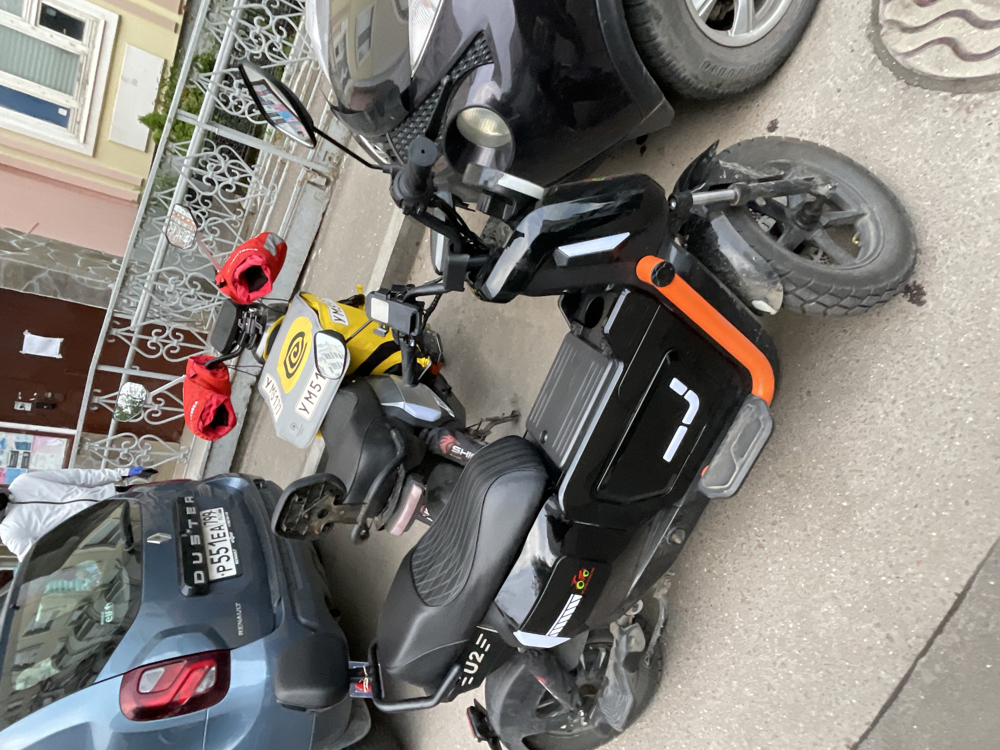 The image depicts a black and orange electric scooter parked on a paved surface in an urban environment. The scooter has a yellow food delivery bag with red delivery boxes attached to its rear. Next to the scooter, there is a blue Renault Duster car with the license plate "P551EA799". The environment suggests an urban setting, which, based on the license plate of the car, could potentially be Moscow, Russia. There are no people clearly visible in the scene, although a partially visible person is on the left of the scooter. The background includes a building with a white window frame and a metal fence.