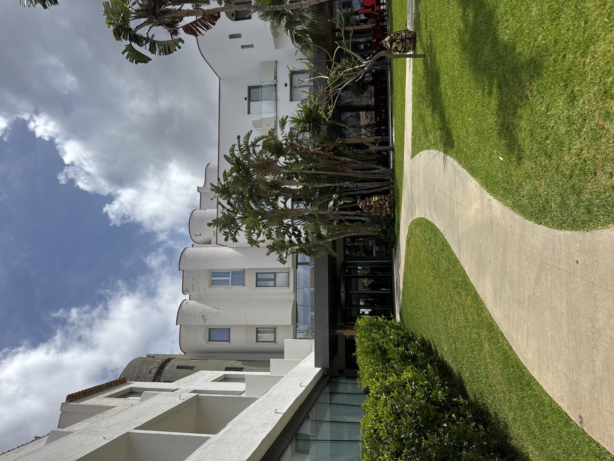 The image depicts a view of the exterior of a hotel building in Isola delle Femmine, Italy. A curved concrete pathway leads through a manicured lawn towards the entrance of the hotel. To the left of the pathway, a dense green hedge lines the edge of the lawn. The hotel itself is a multi-story white building with distinctive architectural features, including rounded sections and multiple windows. Several palm trees and other lush vegetation are visible in front of and around the building. The sky above is partly cloudy. The scene appears to be calm and serene, with no people actively engaged in any visible activities.