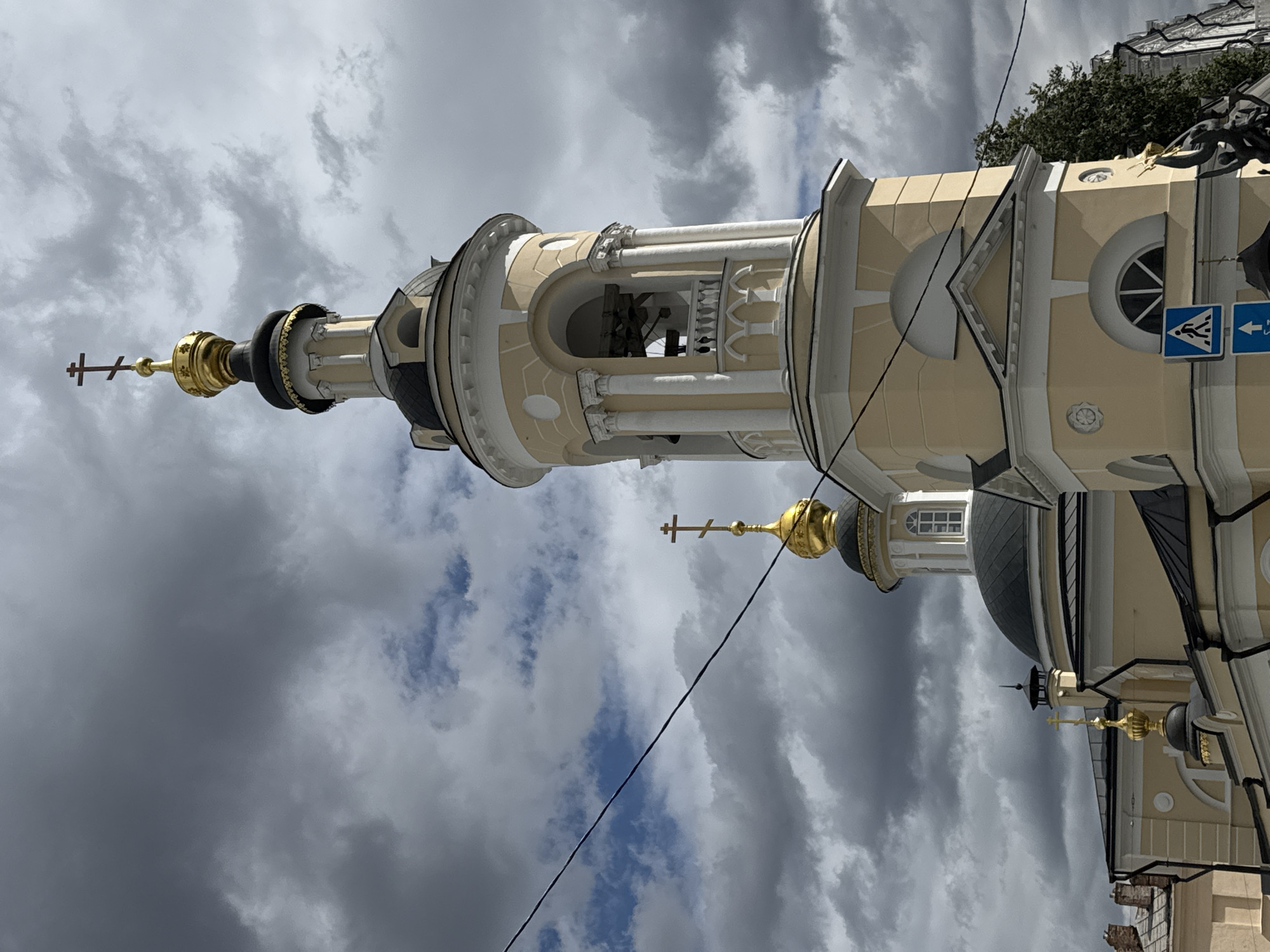 The image shows a close-up view of the bell tower and domes of an Orthodox church, likely in Moscow, Russia, under a dramatic, cloudy sky. The bell tower is tall and slender, painted a pale yellow with white architectural details, including columns and arched openings where bells are visible. Atop the bell tower is a golden dome topped with a multi-barred cross. Further left, another golden dome with a similar cross is visible, belonging to a different section of the church, likely a smaller chapel or part of the main structure. The church itself has a dark, multi-faceted roof and is also painted in pale yellow and white. Power lines are visible crossing the frame horizontally, slightly obscuring the view. In the lower right corner, a dark sculpture of figures and a blue pedestrian crossing sign are partially visible, suggesting the church is located in a populated urban area. The sky is filled with heavy, dark gray clouds interspersed with patches of blue, creating a somber yet striking atmosphere.