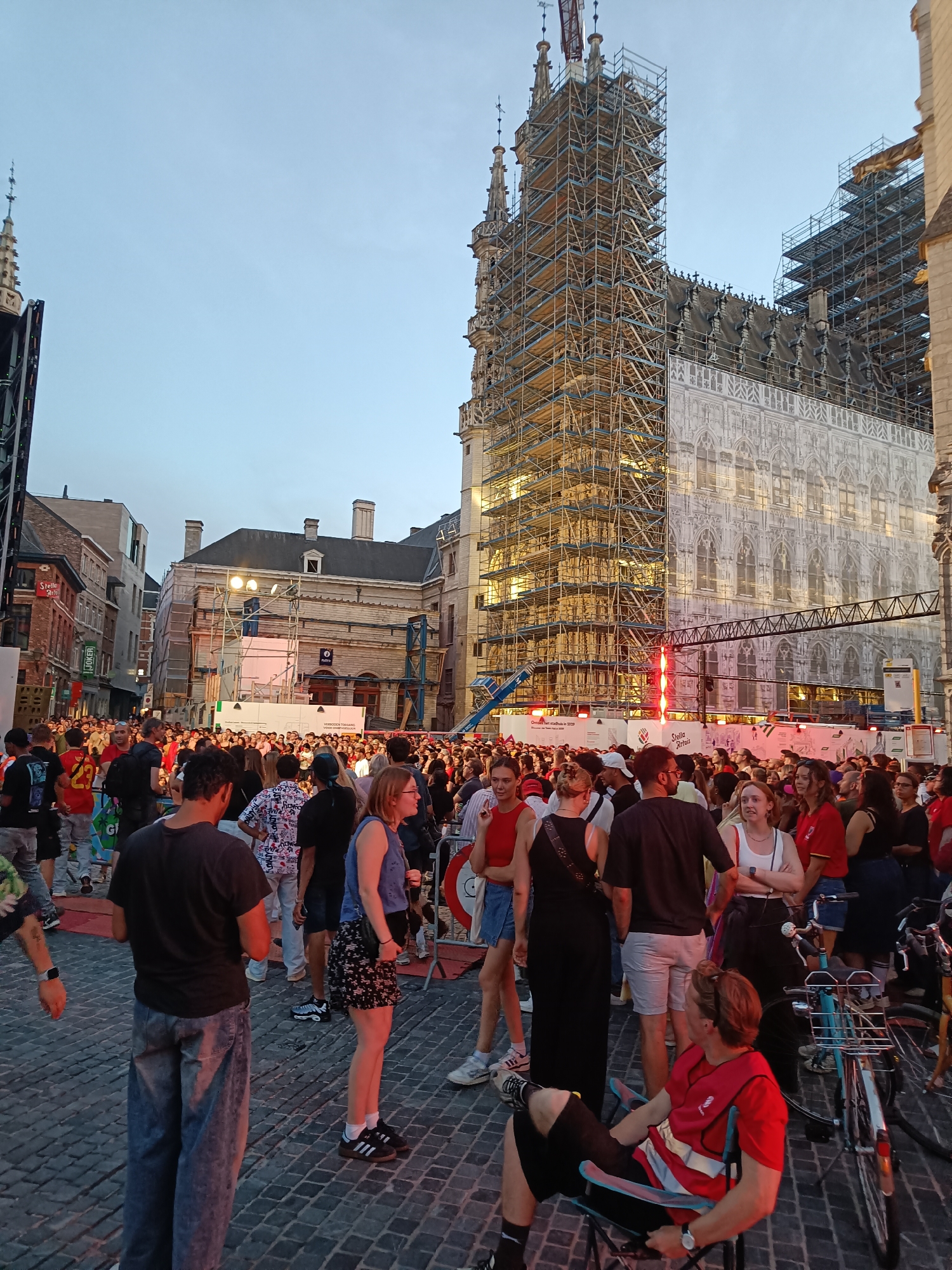 In the city of Leuven, Belgium, a large crowd of people are gathered on a cobblestone street in front of a building undergoing renovation, covered in scaffolding. The building appears to be a historical landmark, possibly a church or a town hall, with ornate architectural details visible beneath a large white banner. The sky is a pale blue, indicating either early morning or late evening. People are dressed in casual attire, with some wearing t-shirts and shorts, suggesting warm weather. Some individuals are standing, while others are sitting on portable chairs. The atmosphere seems festive, with some people engaging in conversation and others looking towards a stage or event area that is not fully visible in the frame. There are barricades set up, potentially to manage the crowd or delineate an event space. Several bicycles are parked nearby. The scene captures a moment of public gathering in a European city.