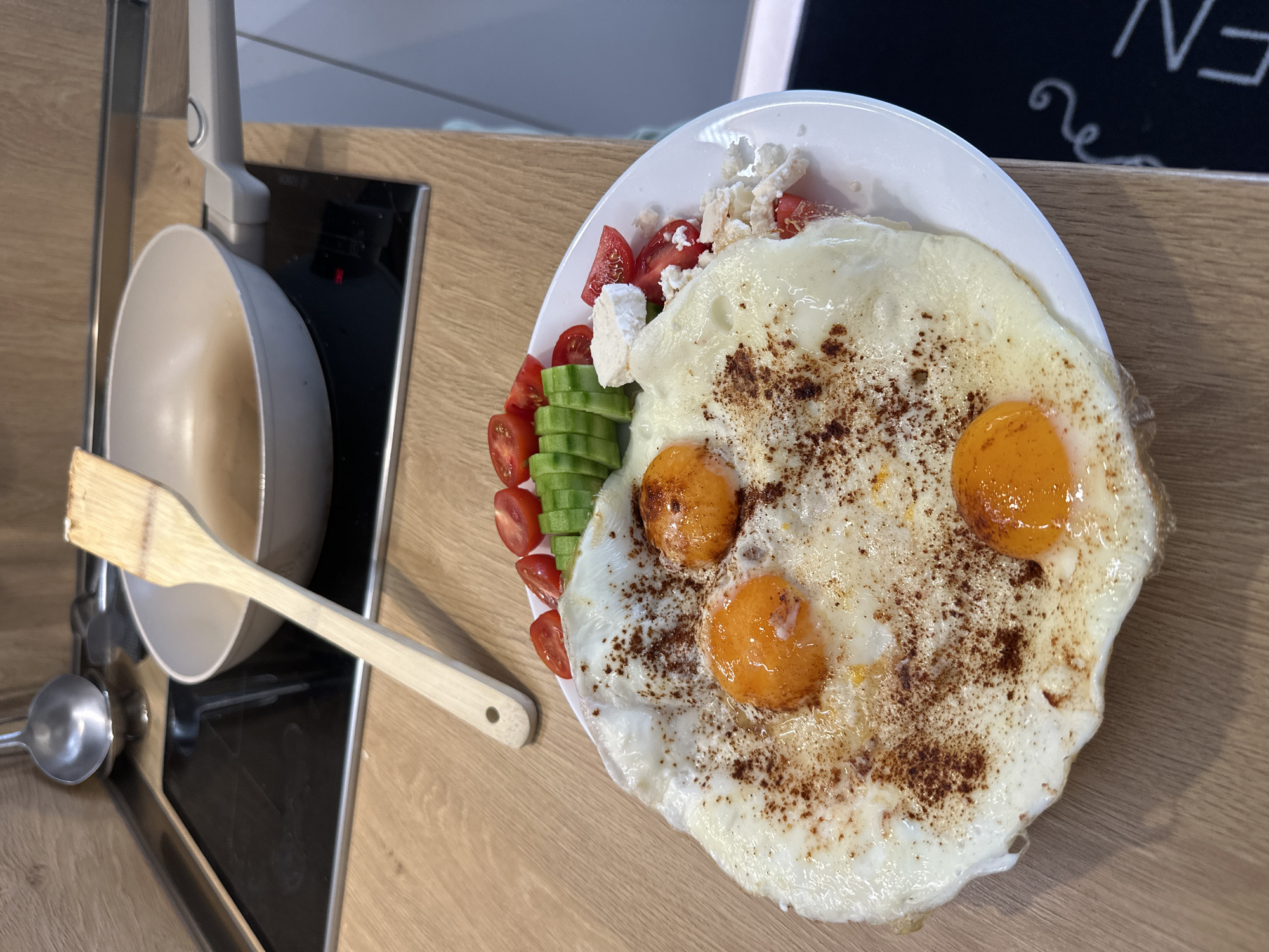A high-angle shot captures a freshly prepared meal, likely breakfast or brunch, featuring three fried eggs on a white plate. The eggs have distinct runny yolks and slightly crispy whites, and are sprinkled with a dark reddish-brown spice, possibly paprika or sumac. Accompanying the eggs are vibrant sides of sliced cherry tomatoes, neatly cut cucumber slices, and crumbled white cheese, which, in the context of Plovdiv, Bulgaria, is likely feta or Sirene cheese.
The meal is presented on a light wooden countertop, suggesting a kitchen or dining area. To the left of the plate, a light-colored frying pan with a grey handle sits on a black induction hob, indicating recent cooking. A wooden spatula rests on the countertop, partially overlapping the pan, and a metal ladle is visible further to the left. In the upper right corner, a partial view of a black surface with white, hand-drawn text and a decorative swirl is visible, showing fragments that look like "N" and "E," possibly from a chalkboard sign. The bright illumination suggests a daytime setting. The scene conveys a sense of a home-cooked, nutritious, and appealing meal.