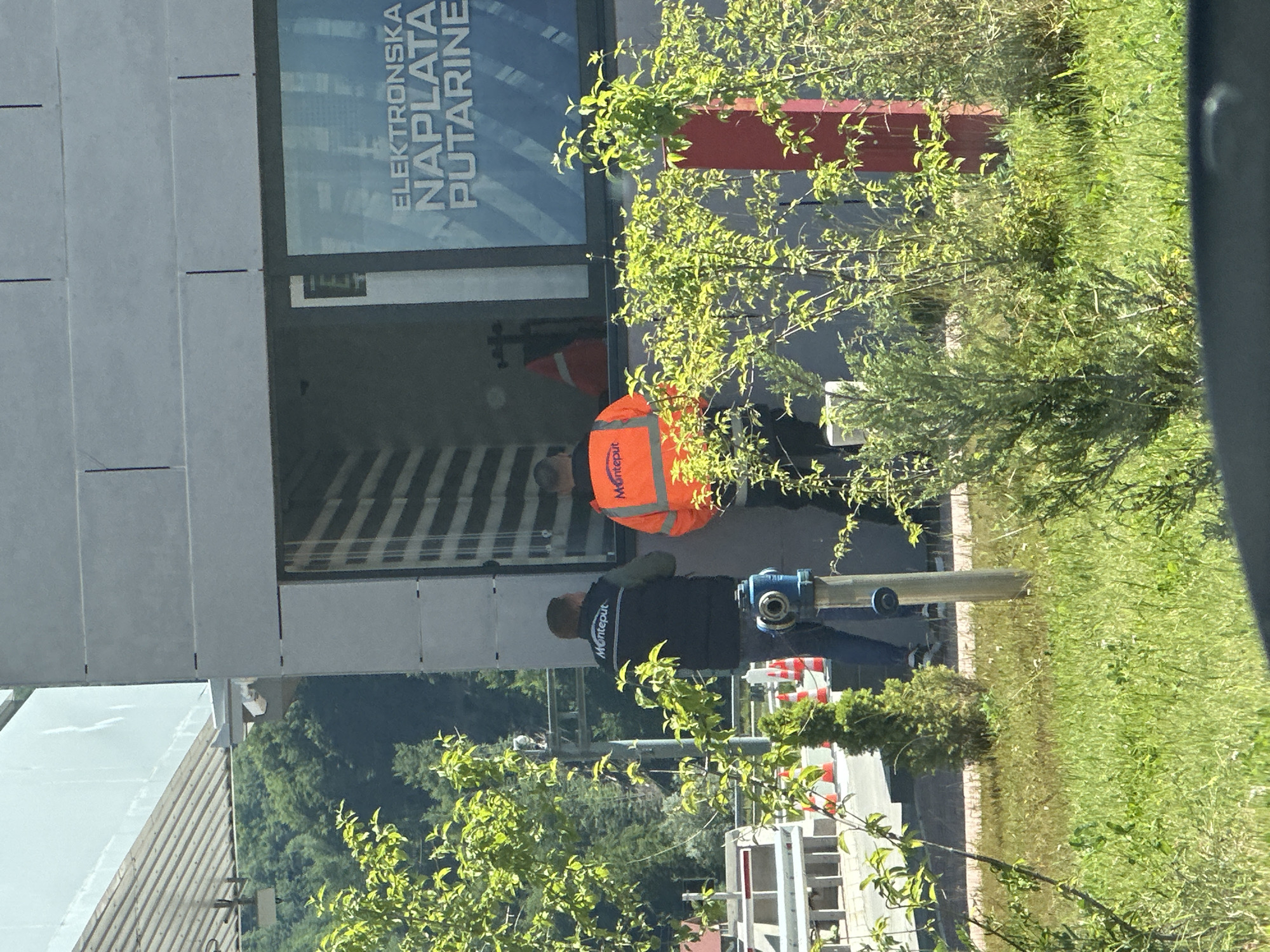 In Klopot, Montenegro, two male individuals, likely employees, are present near a toll booth. One man, wearing a dark vest with "Monteput" branding, stands facing away from the camera. The other man, in an orange reflective vest, also facing away, has "Monteput" visible on his clothing. They appear to be engaged in some activity related to the booth, which has signage that reads "ELEKTRONSKA NAPLATA PUTARINE." The environment surrounding the booth includes lush green vegetation, a blue fire hydrant, and a red post. The overall scene suggests an operational toll collection point.