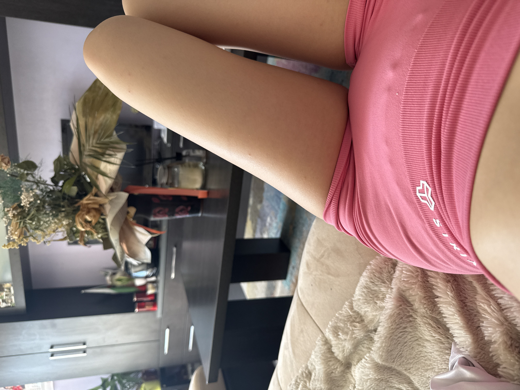 The image shows a close-up of a person's legs, wearing pink athletic shorts, while lying on a sofa. The background is a living room with a coffee table, a television, and a cabinet with decorative dried flowers. The location is identified as Sofia, Bulgaria.