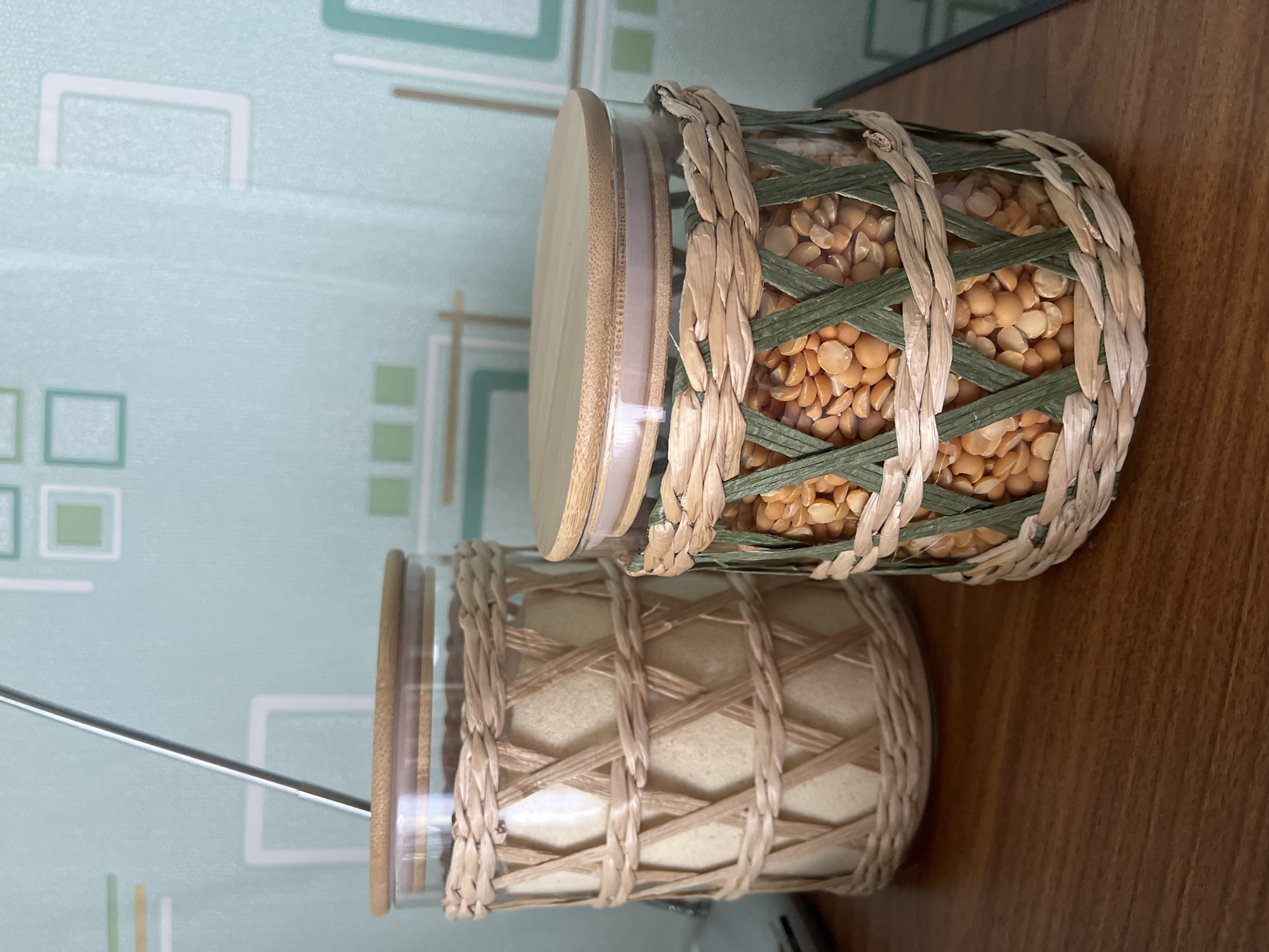 This media file features two glass jars with woven casings and bamboo lids, placed on a wooden surface. The top jar is filled with yellow split peas, and its woven casing is a mix of natural straw and green reeds. The bottom jar contains a light-colored granular substance, possibly sugar or flour, and its woven casing is entirely made of natural straw. Both jars have clear glass bodies and are topped with natural bamboo lids. The background shows a wall with a light blue geometric pattern. The lighting suggests an indoor setting, possibly a kitchen or pantry, with no indication of the time of day or weather. There is no visible text in the image. The overall scene is static, focusing on the decorative storage jars.