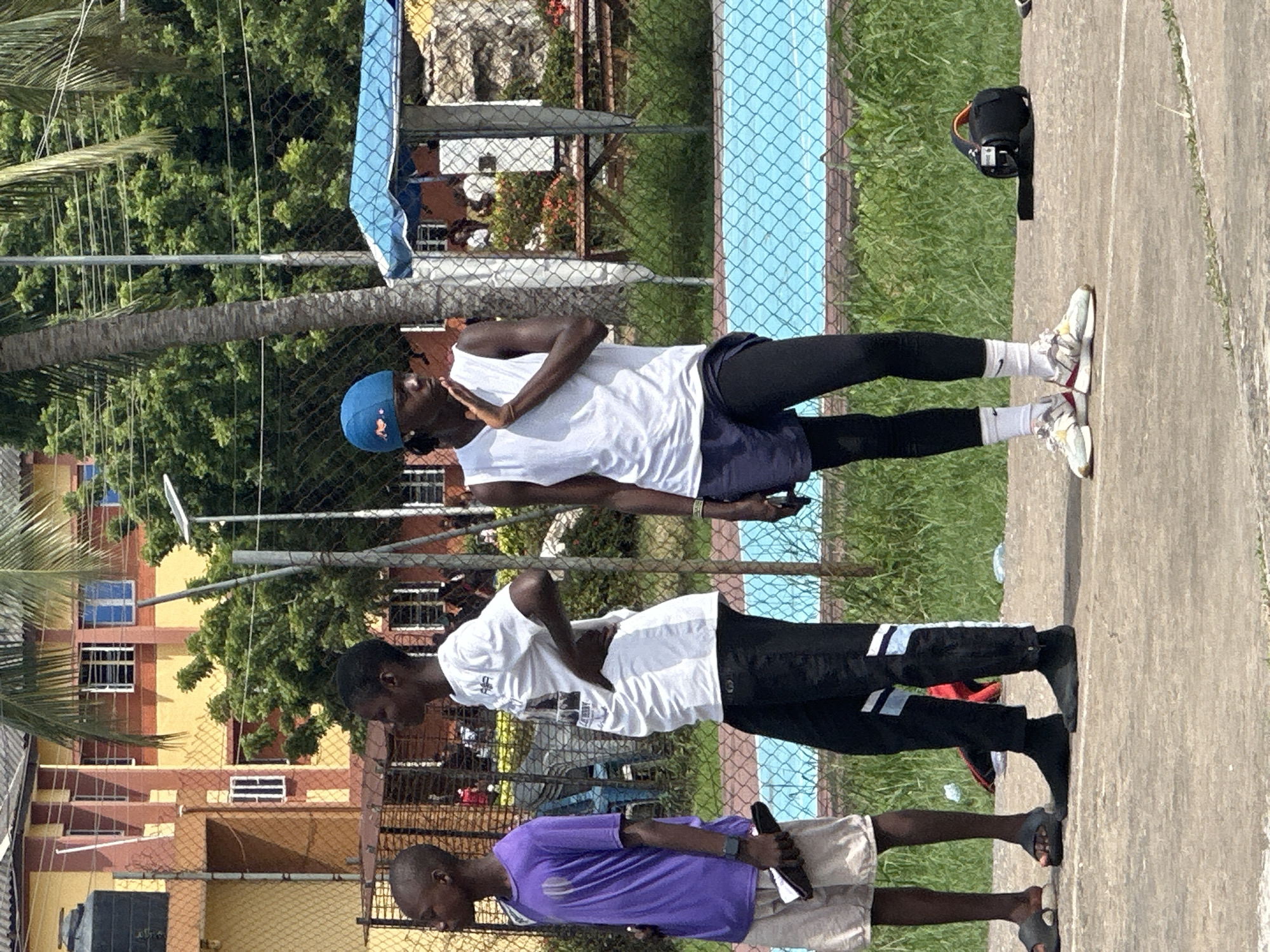 The image captures three young males standing on a paved surface, possibly an outdoor court or track, in what appears to be a recreational area within the city of Ajara, Nigeria. The background features a building with a yellow and red facade, surrounded by lush green trees and a palm tree. A blue net fence separates the court from a bright blue swimming pool area.
On the left, a boy in a purple t-shirt and light-colored shorts holds a black object in his hand. Next to him, a second boy stands with his hands on his hips, wearing a white t-shirt and black pants with white and blue stripes. The third and tallest individual, on the right, wears a blue cap, a white sleeveless shirt, and dark shorts over black leggings. He has his right hand raised to his chin, with his mouth slightly open. His posture suggests he might be speaking or listening intently.
Other elements visible include a black bag with an orange handle on the ground near the third individual, a metal fence, and some discarded items on the ground. The overall scene is bright and appears to be a daytime outdoor activity.