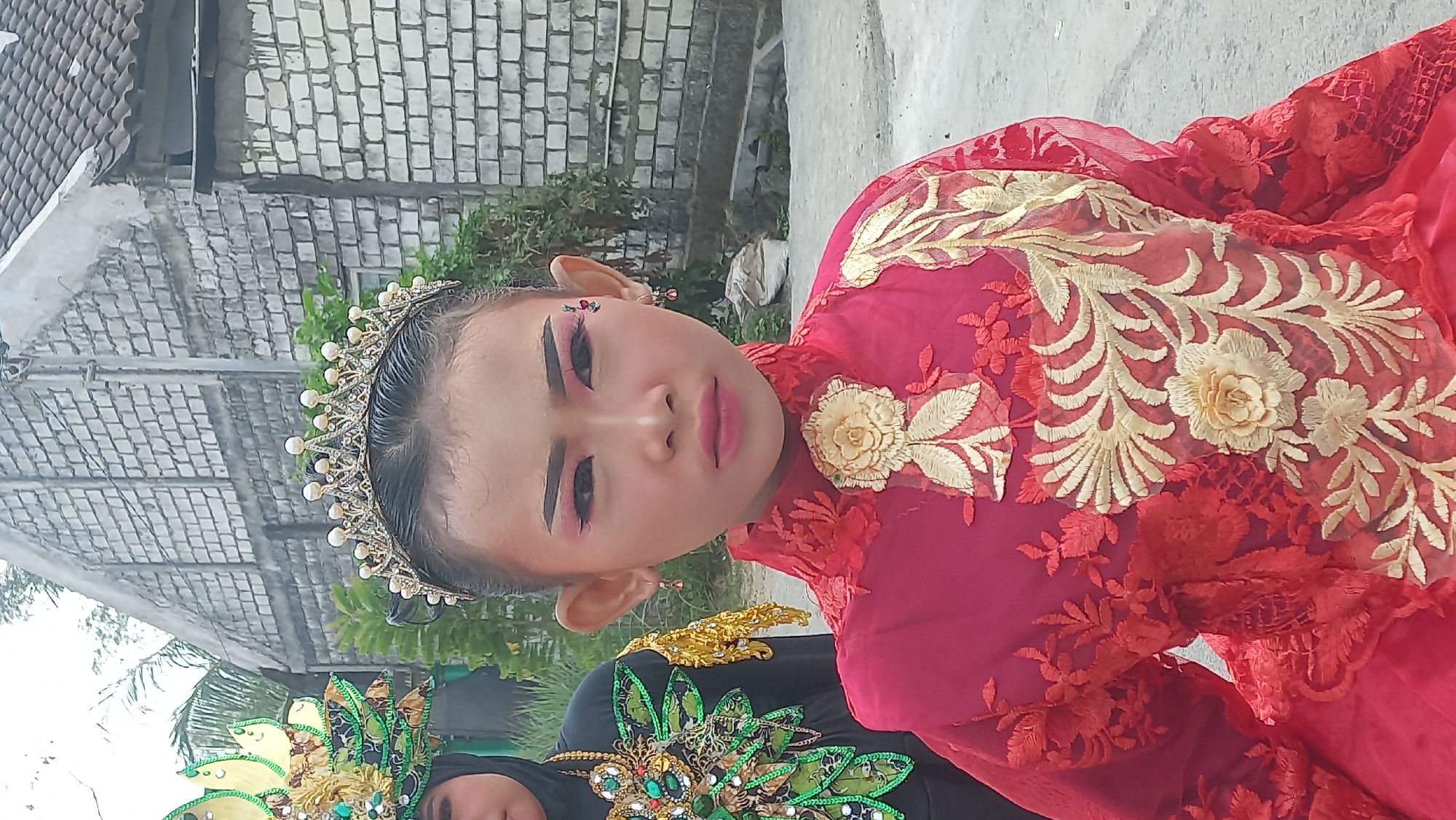 A young Indonesian girl, adorned in a red and gold ornate costume with a pearl-studded tiara, stands in the foreground of an outdoor scene. Her face is made up with dramatic pink eyeshadow and meticulously shaped eyebrows. A second, partially visible individual in a similar elaborate costume is to her left. The background consists of a weathered brick building with a tiled roof. The setting appears to be a street in the village of Kedungmegarih, Indonesia, indicated by the architectural style and the presence of lush greenery. The lighting suggests it is daytime.
