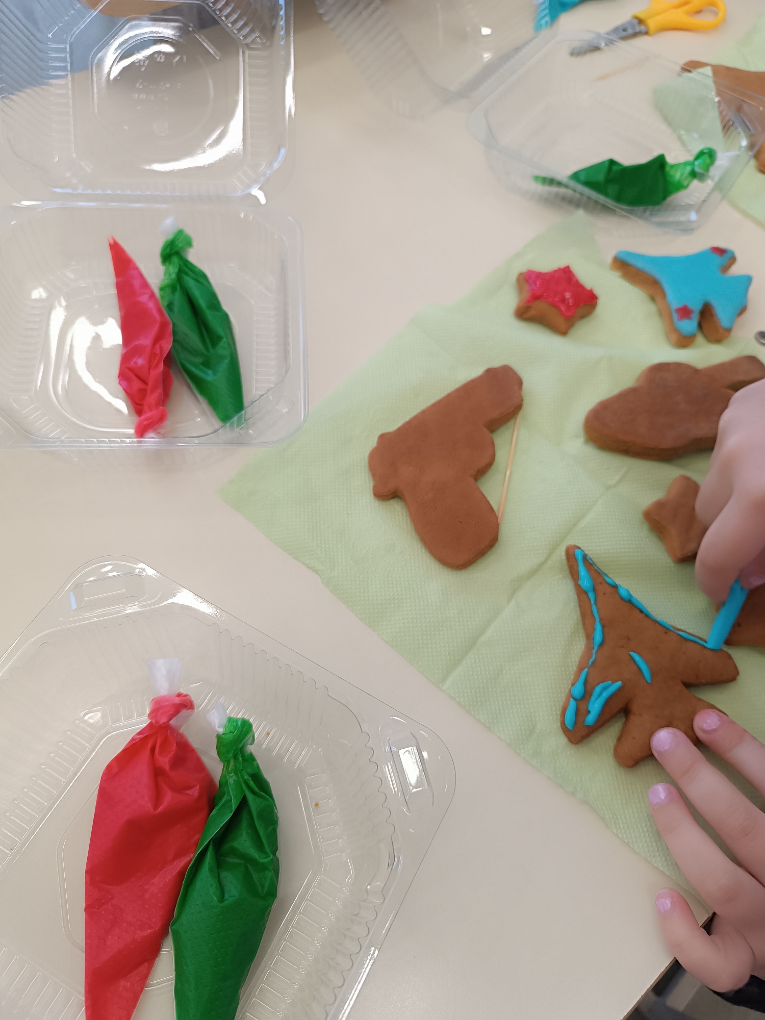 A high-angle shot captures a table setting with various items suggestive of a baking activity, possibly in a domestic or educational setting. Two clear plastic containers sit on the left side of the frame, each holding a pair of icing bags filled with red and green icing, respectively. To the right, on a light green napkin, several gingerbread cookies of different shapes, including a star, an airplane, a gun, and an abstract form, are partially decorated with blue and red icing. A child's hand, with nails painted in a pale purple hue, is actively decorating the gingerbread tree cookie with blue icing. The environment includes a light-colored tabletop and additional clear plastic containers in the background, with one containing green icing, implying an ongoing or prepared cookie-decorating project.