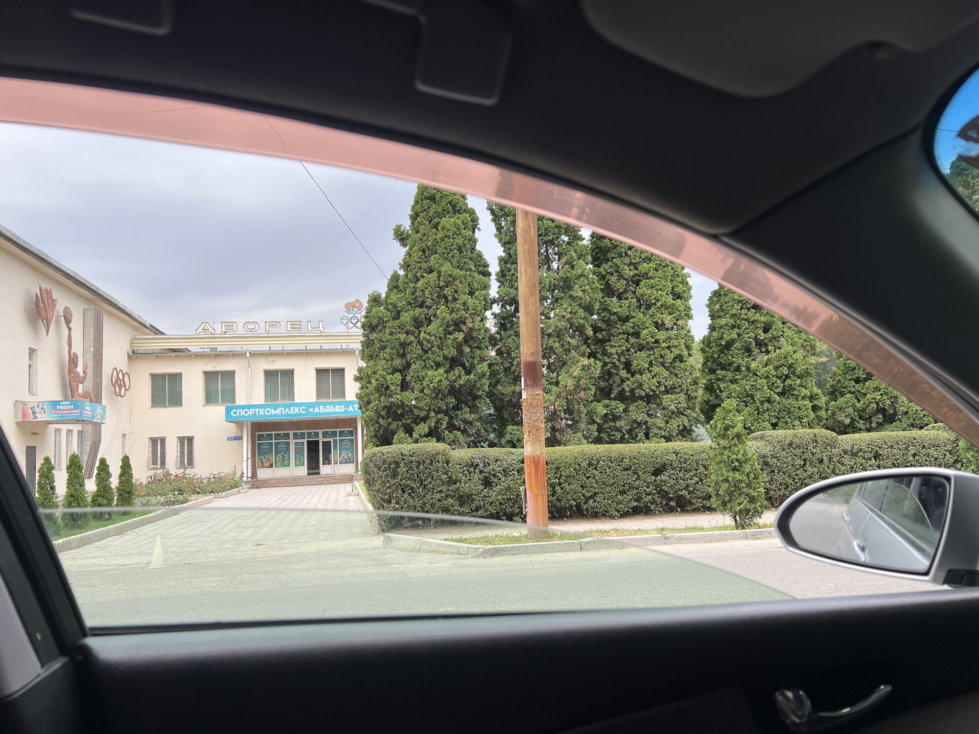 The image shows a view from inside a car, looking out of a partially open window. The scene outside is a building with a sign that reads "СПОРТКОМПЛЕКС «АБДЫШ-АТА»" (Sports Complex "Abdysh-Ata"). The building appears to be a sports facility, with architectural elements suggesting its purpose, such as a depiction of athletes on the left side. In front of the building, there is a pathway leading to the entrance, flanked by manicured bushes and trees. Several cypress trees stand tall, with a rustic wooden pole in the foreground. The sky is overcast, indicating a cloudy day. The reflection in the car's side mirror shows another vehicle. The overall impression is that of a daytime scene in a location that appears to be a sports complex in Kant, Kyrgyzstan, possibly on a day when activities are not in full swing, given the quiet and somewhat overcast atmosphere.