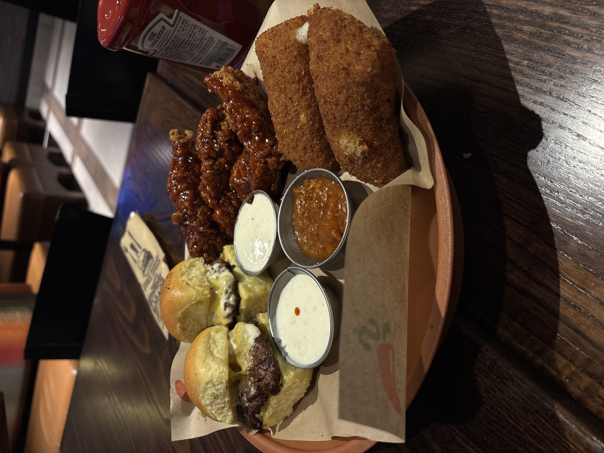 This image shows a plate of appetizers on a wooden table. The appetizers include chicken tenders with a dark glaze, fried mozzarella sticks, sliders, and three small metal cups filled with different sauces: white sauce with a red dot, white sauce, and an orange sauce. A bottle of ketchup is visible in the background, partially obscuring the chicken tenders. The food is arranged on a terracotta-colored plate lined with brown paper. The photo may be taken in Puebla, Mexico.