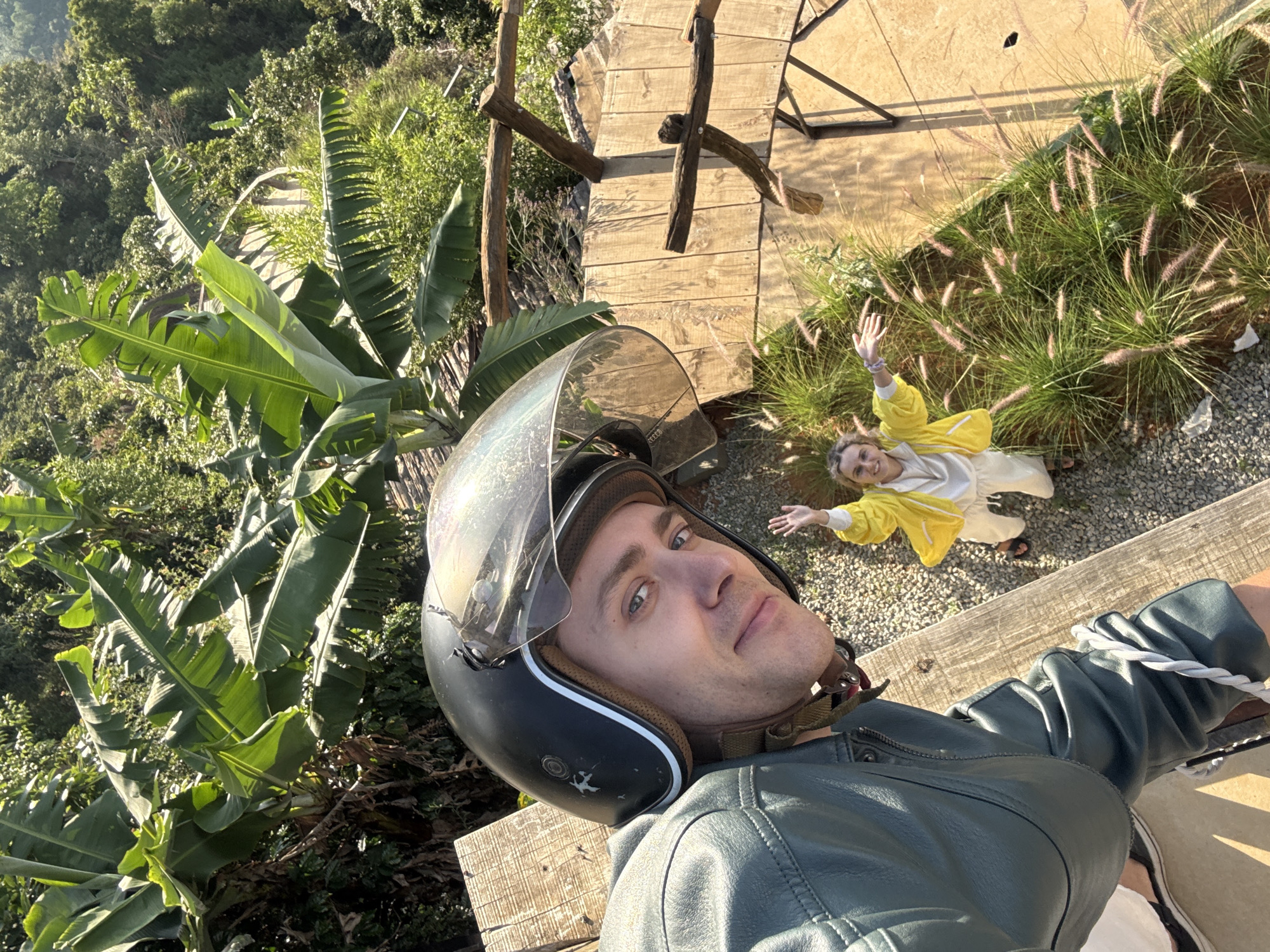 A high-angle shot depicts a man in a motorcycle helmet taking a selfie. The man is wearing a green leather jacket and the helmet is black with a clear visor. A woman, dressed in a yellow jacket and white pants, lies on the ground nearby, waving both arms in the air. In the background are lush green banana trees, other trees, and tall grass. A wooden structure with planks is partially visible. The scene is located in Phát Chi, Vietnam.