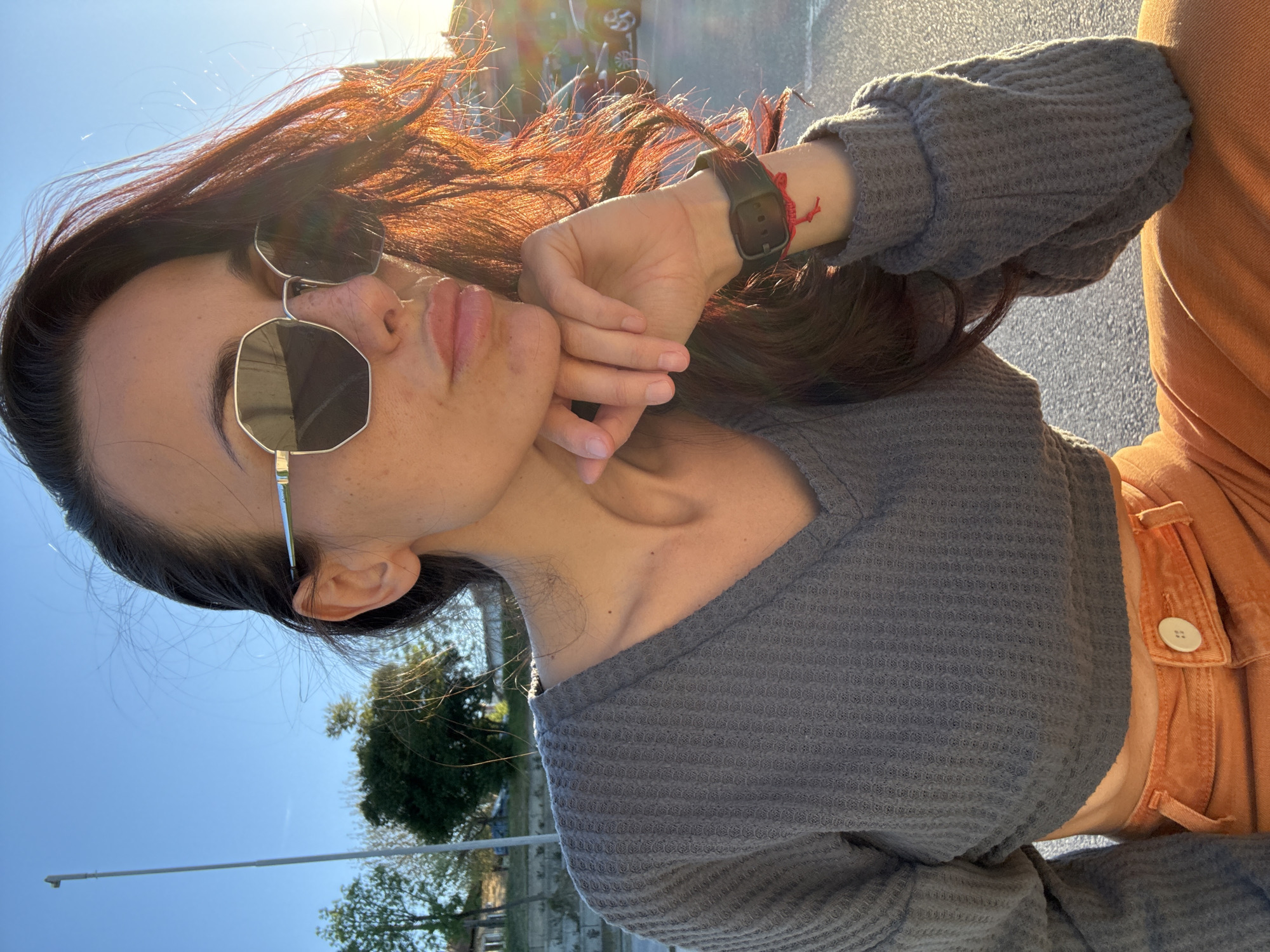A medium shot, taken from a low angle, features a young woman outdoors under bright, warm sunlight, likely in Pleven, Bulgaria. The image is rotated 90 degrees, showing the woman's profile from the left side. She has dark, shoulder-length hair, which catches the strong backlight, creating reddish highlights and a noticeable lens flare. She is wearing octagonal-shaped sunglasses with dark lenses and silver frames, partially obscuring her eyes. Her full lips are visible.
She is dressed in a grey, long-sleeved, v-neck top with a waffle-knit texture. On her left wrist, she wears a black digital watch and a red string bracelet. Her right hand is gently placed near her jawline. The lower right portion of the image shows a hint of orange or rust-colored high-waisted pants or a skirt with a visible white button.
The background is softly blurred but reveals a clear blue sky, a few green trees, and what appears to be asphalt ground. A thin, white pole or pipe is visible against the sky. The intense, warm light and lens flare suggest it is late afternoon or golden hour on a clear day. The woman maintains a neutral and somewhat pensive expression while posing for the camera. There is no visible text in the image.