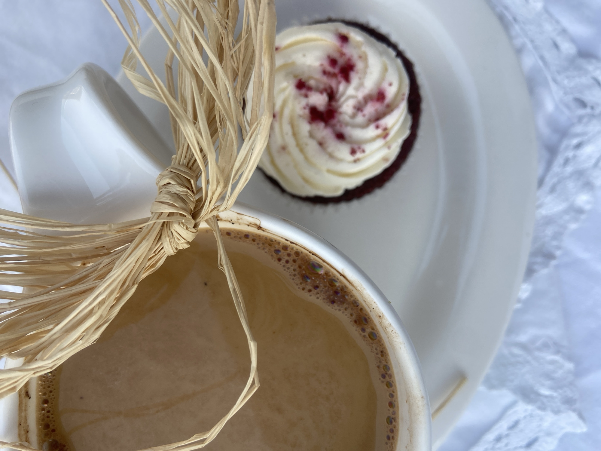A top-down, close-up view captures a coffee cup, a cupcake on a white plate, and a bundle of dried straw. The coffee cup, filled with a light brown beverage and a rim of bubbles, occupies the bottom left of the frame. A bundle of tan straw is tied around the cup's side. To the upper right, a cupcake with white frosting and a dusting of red crumbs rests on a shiny white plate. The scene unfolds against a backdrop of textured white fabric. The image does not contain information that could identify the location as Budva, Montenegro.