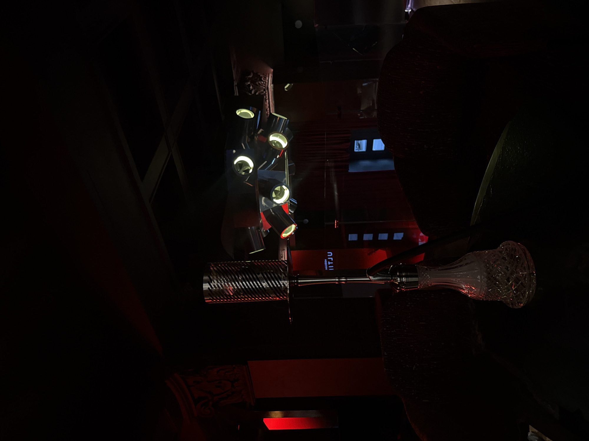 The image displays a dimly lit indoor scene, likely a sophisticated bar, restaurant, or lounge, characterized by deep shadows and artificial light sources. The perspective appears to be upward or sideways, focusing on architectural and decorative elements.
On the left, a cluster of modern, ceiling-mounted spotlights illuminates the space with a warm, yellowish glow. Their metallic casings reflect the ambient light. To the right, a prominent, ornate glass object, possibly a decanter or a part of a decorative fixture like a hookah, is visible. It features intricate faceted patterns and reflects a reddish hue from surrounding elements. Adjacent to it, a metallic, cylindrical object with a ribbed, spiraled texture adds another decorative touch.
In the background, a rectangular panel or sign in a vibrant red color displays white text, which clearly reads "HITJU" and partially "MADISON" above it. Further back, above this red panel, several small, square shapes suggest windows or decorative cutouts, revealing a faint, lighter blue or grey tone behind them. Hints of dark, possibly upholstered or carved architectural features, including a subtle reddish pattern at the bottom left, contribute to the venue's elegant and somewhat mysterious ambiance. The overall impression is one of a stylish evening setting, with no people visible.