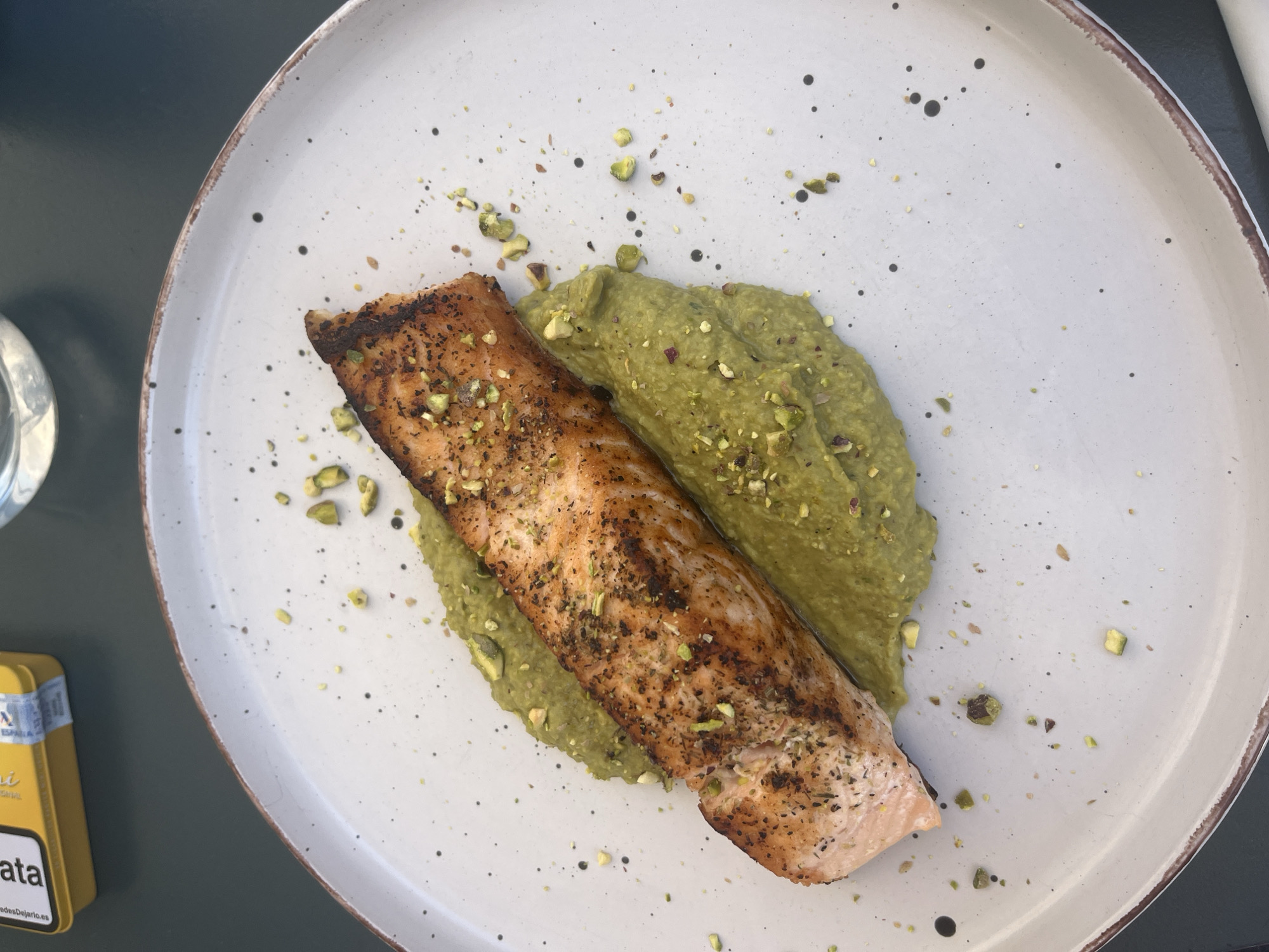 Overhead shot of a white speckled plate featuring a piece of grilled salmon resting on a bed of green purée, likely pea or pistachio. Chopped pistachios are scattered around the plate as garnish. The plate sits on a dark green table, partially revealing a yellow cigarette pack branded “Ata” and a glass. The photo was likely taken outdoors, in the city of Alicante/Alacant, Spain.