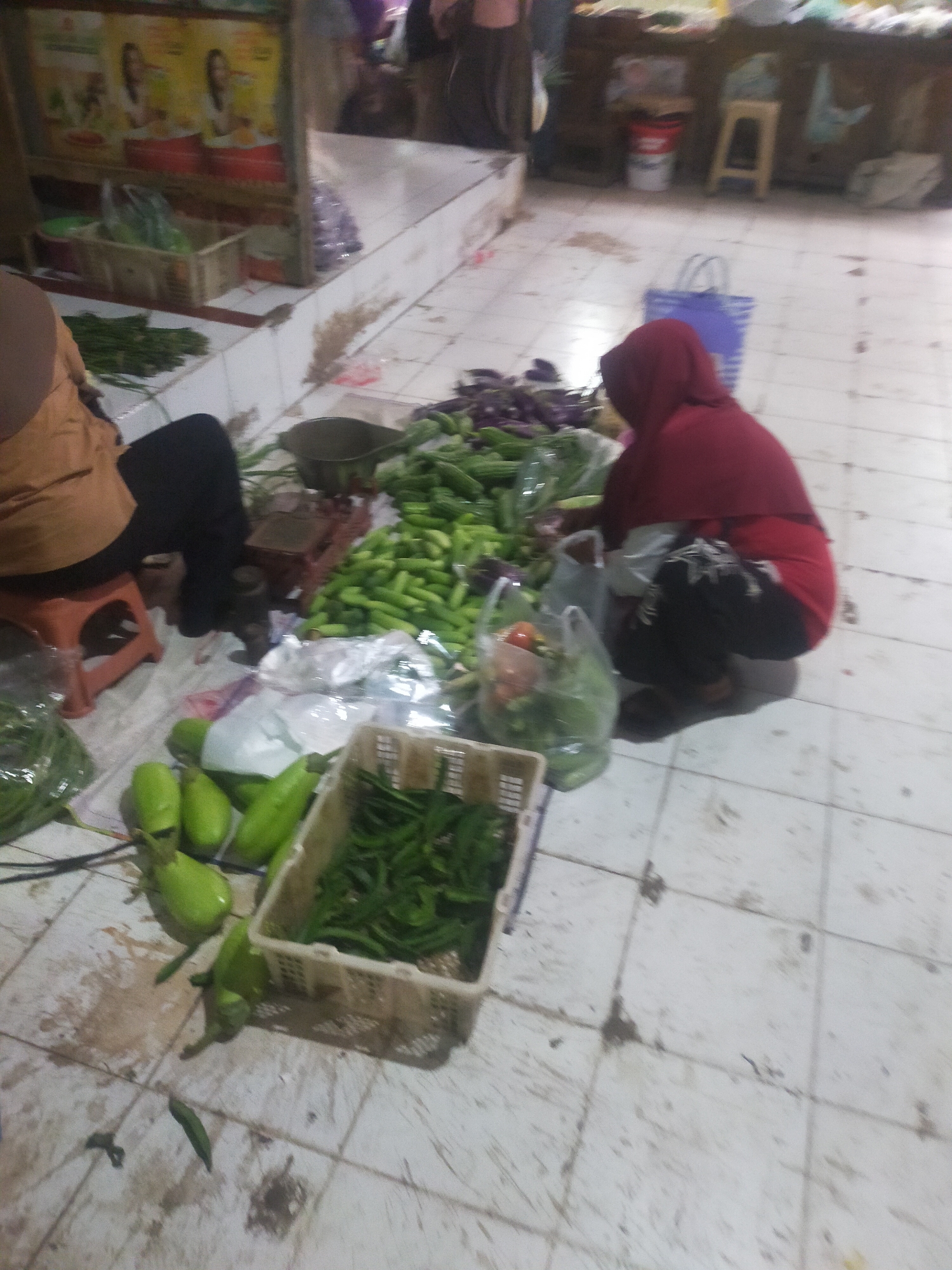 In Trayeman, Indonesia, a market scene unfolds with a focus on fresh produce. The foreground is dominated by an assortment of green vegetables, including what appear to be eggplants and long beans, some arranged in plastic baskets and others laid out on the tiled floor. A person wearing a red hijab and dark clothing is crouched down, seemingly engaged with the produce. To the left, another individual in a brown shirt and black pants sits on an orange stool, partially visible. In the background, market stalls are suggested by shelving and displays, with more vegetables and what might be packaged goods. The environment appears to be an open-air or covered market, with tiled flooring that shows some signs of wear and dirt. Bags, both plastic and fabric, are scattered around, indicating shopping activity.