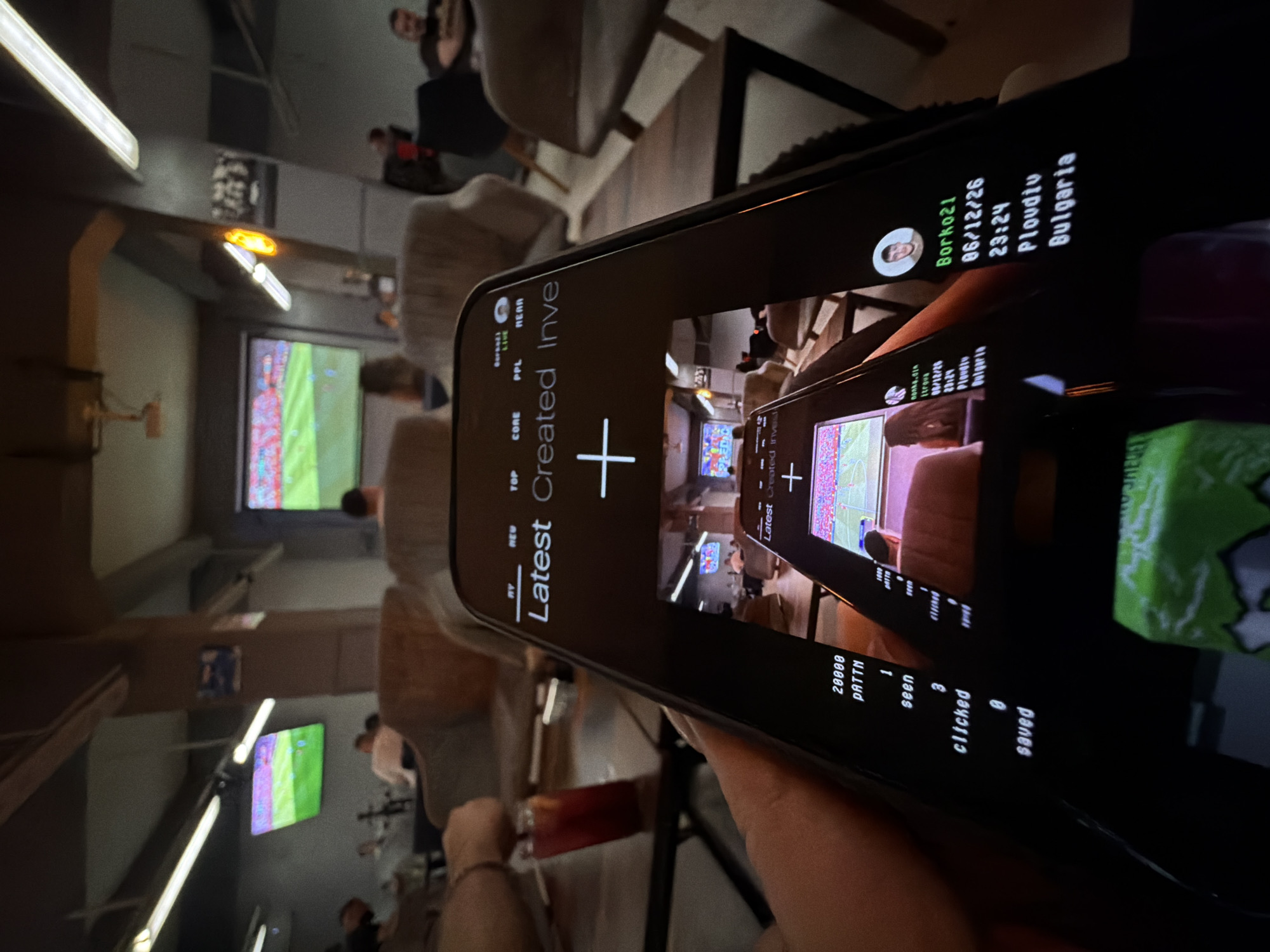 The image shows a person holding a smartphone displaying a user interface with text and images. The phone screen shows the text "Latest Created Inve" with a plus sign. Below this, a smaller image within the phone screen shows a similar interface with text that reads "20000 PATTN," "1 seen," "3 clicked," and "0 saved." Further down the phone screen, there is information including a profile picture, the name "Borko21," a date "06/12/26," a time "23:24," and the location "Plovdiv, Bulgaria."
The background suggests an indoor setting, possibly a cafe or lounge, with people visible in the distance and screens displaying what appears to be a soccer match. There are tables and chairs in the environment. The overall scene is dimly lit. The presence of the "Plovdiv, Bulgaria" text on the phone screen indicates the location.
