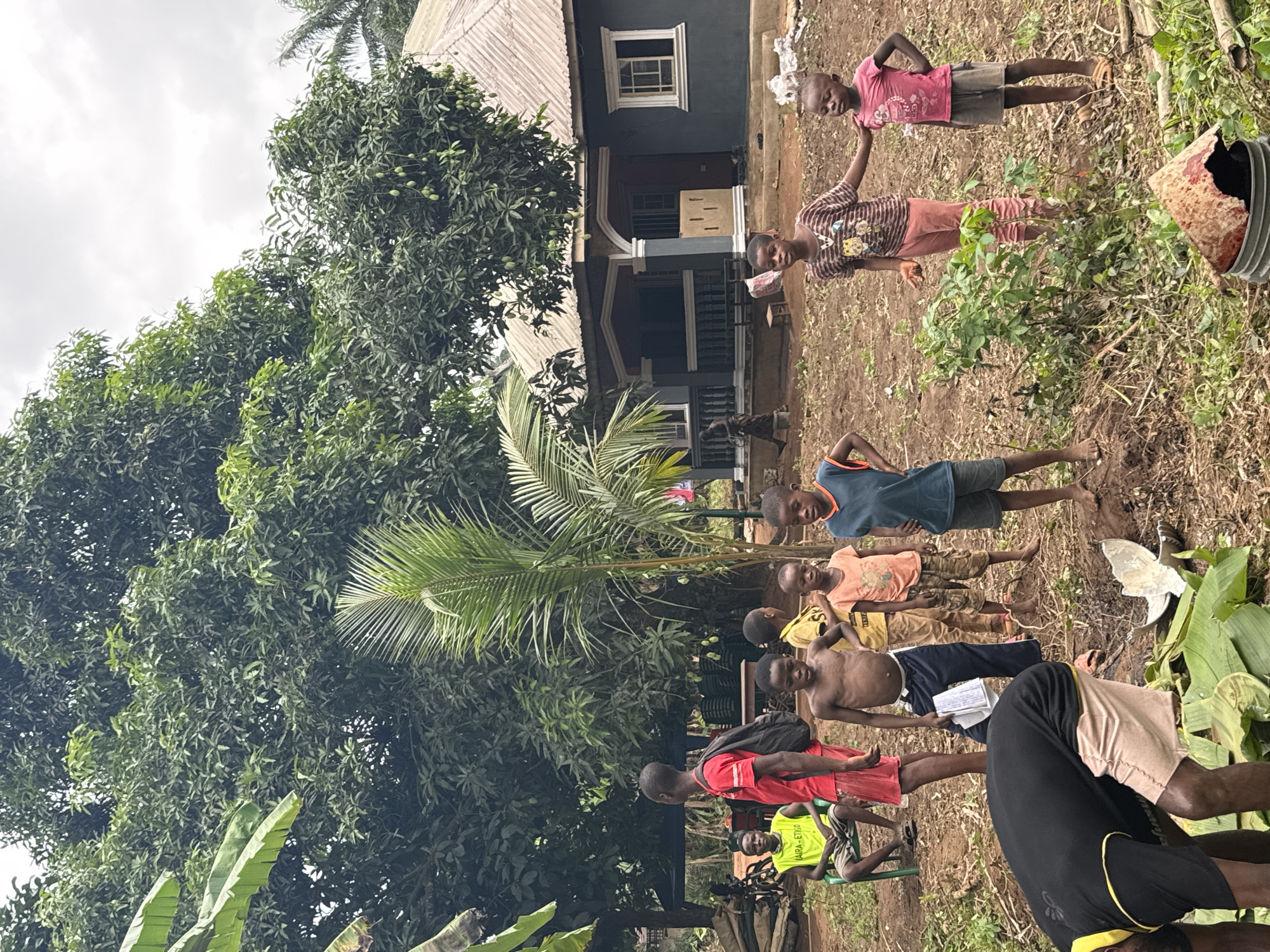 The image depicts a group of children in Eziudo, Nigeria, standing outdoors in front of a house. The house is two-storied, painted dark gray with white trim, and has a balcony. A large leafy tree is beside the house, and there is a palm tree in the foreground. The ground is a mix of dirt and sparse vegetation. The children are of varying ages, dressed casually, some barefoot. One is shirtless and holding a book. Another is seated on a plastic chair.