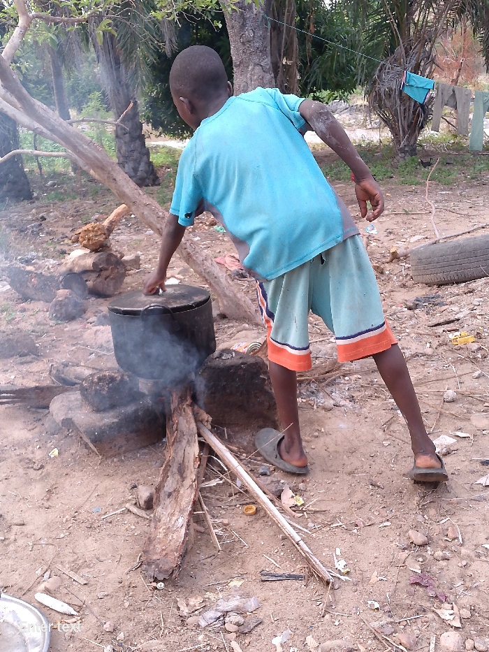 A young person stands over a makeshift cooking fire in an outdoor location. The individual wears a short-sleeved blue shirt, shorts with orange trim, and sandals. They are reaching towards a large, black pot resting on the fire, which is constructed of logs, rocks, and a tire. Smoke rises from the fire. The ground is dirt and littered with debris. Trees and laundry hanging on a line are visible in the background. The location appears to be in an underdeveloped area.