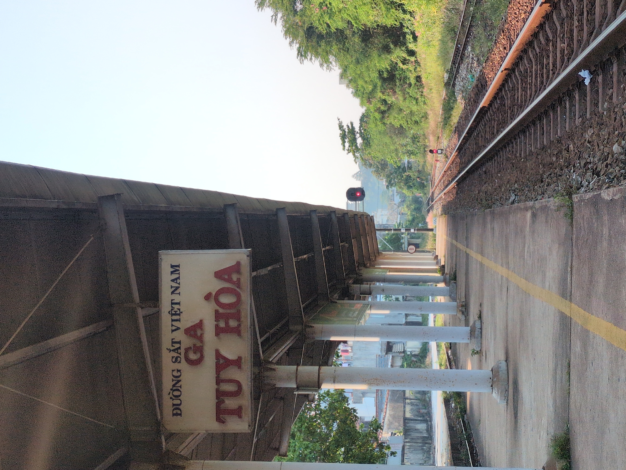 The image shows a view of Tuy Hòa train station in Vietnam. A platform covered by a metal canopy and supported by columns runs parallel to a railway track. The station's sign displays the name "TUY HÒA" in large red letters and "ĐƯỜNG SẮT VIỆT NAM" in smaller dark letters above it. A yellow line marks the edge of the platform. A red signal light is visible further down the track, indicating a stop. The environment features vegetation alongside the tracks. The sky is visible above the trees and the station roof, appearing hazy.