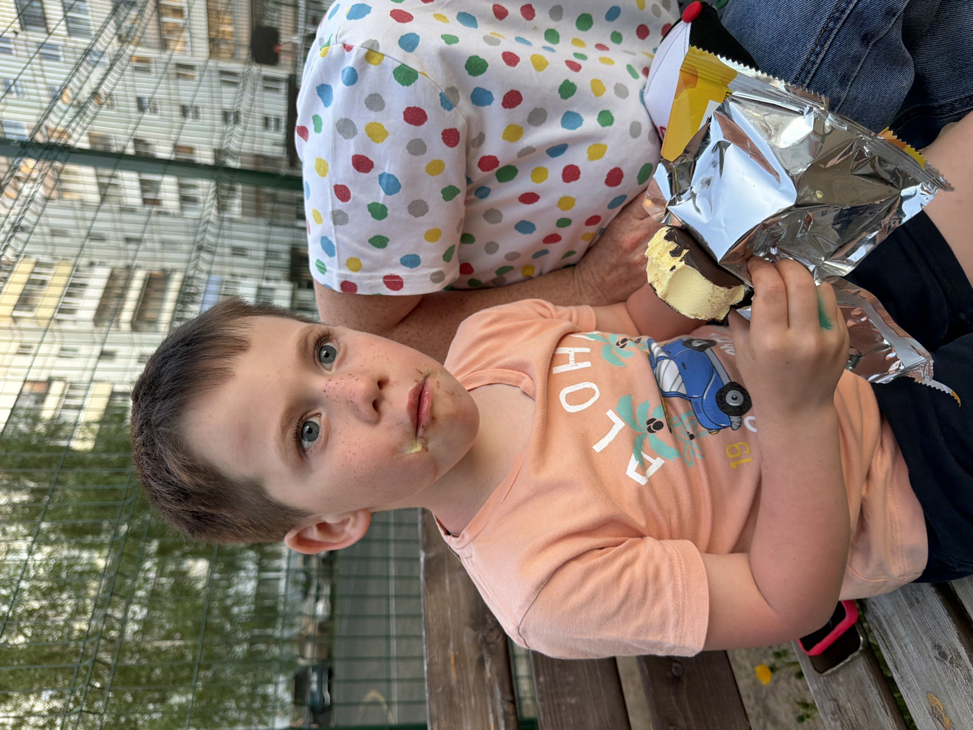 A close-up, medium shot depicts a young boy with chocolate and ice cream smeared around his mouth. He is fair-skinned with short brown hair and blue eyes, freckles, and wearing a light orange t-shirt printed with the word "ALOHA," a blue car, and palm trees. He is sitting on a wooden bench next to an adult wearing a white shirt with multi-colored polka dots and denim jeans. The boy is holding a half-eaten ice cream sandwich in a shiny foil wrapper. The setting appears to be outdoors, possibly a park or recreational area, with a green metal fence visible in the background and the blurred image of a multi-story building. The location is Chaikovsky, Russia. A cell phone is visible on the bench.