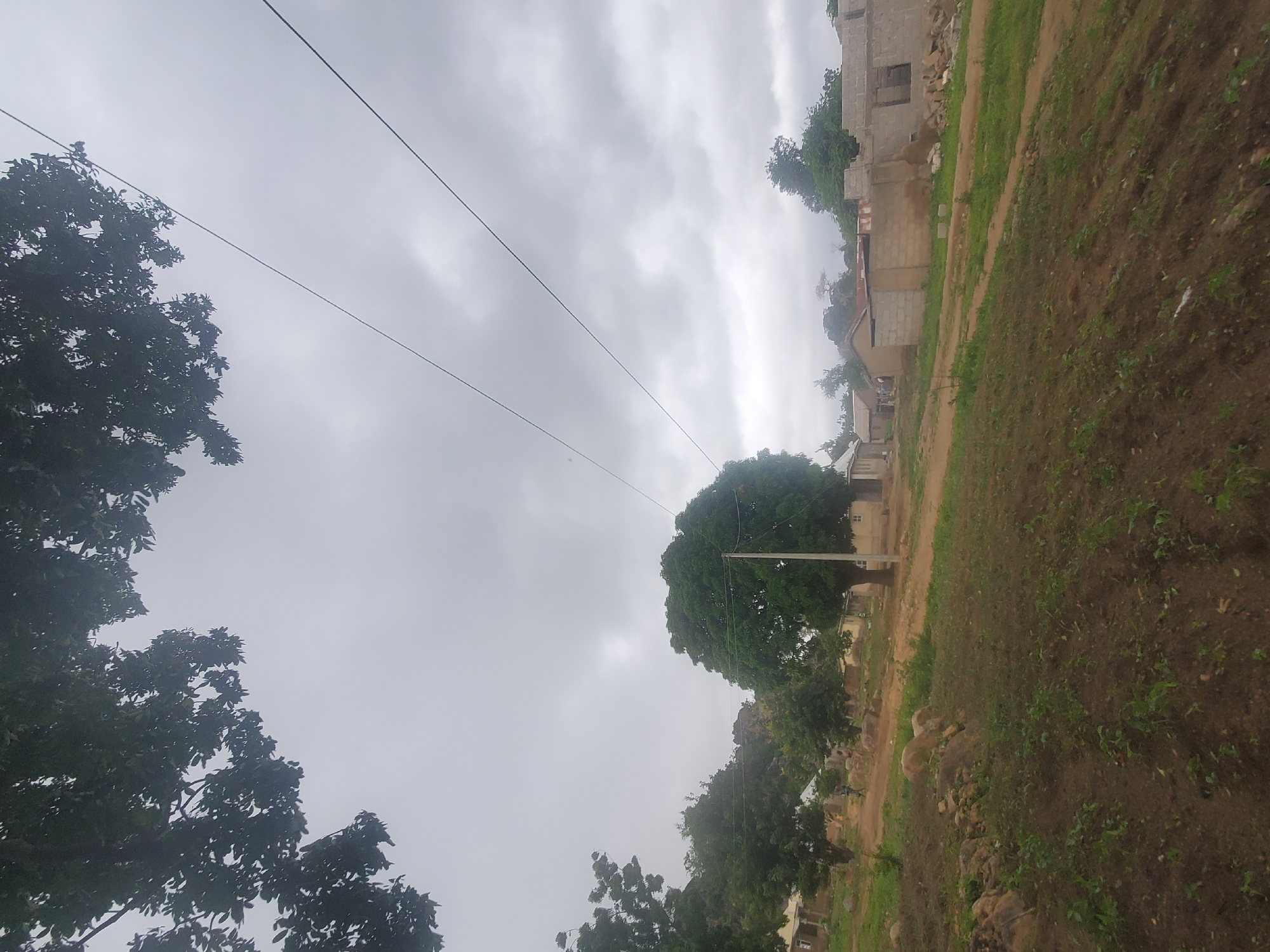 The image depicts a view of a dusty, unpaved road leading through a rural settlement in Kabwir, Nigeria. The sky is overcast with thick clouds. On the left, there are trees and some low-lying buildings. A prominent large, leafy tree stands in the center of the frame. To the right, more buildings are visible, some of which appear to be under construction, with exposed concrete blocks. Power lines are strung across the upper portion of the image. The foreground shows a sloped area with sparse vegetation and some rocks. A figure, possibly a person, is faintly visible walking on the road in the middle ground.