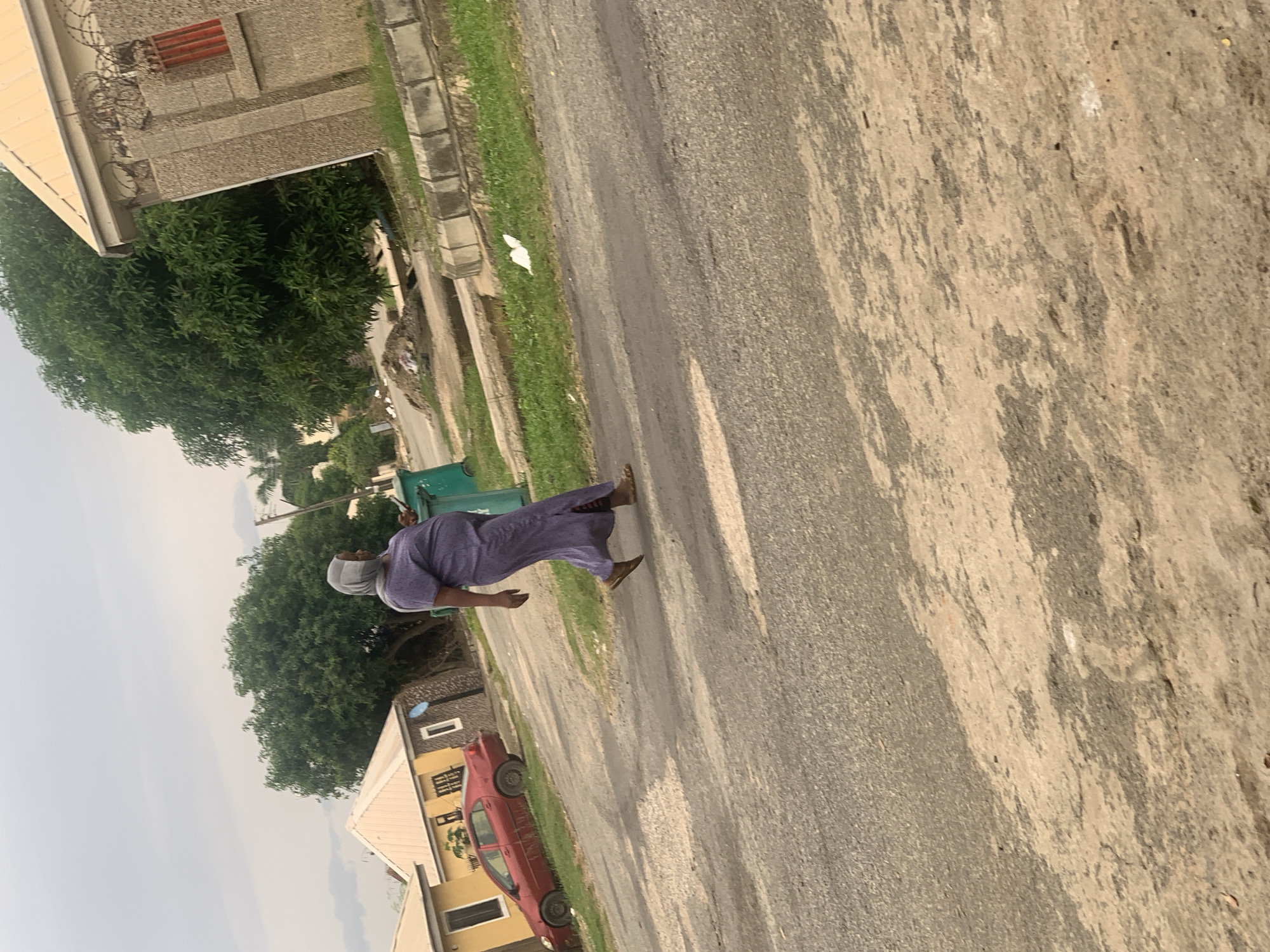 An outdoor, ground-level, medium shot captures a woman wearing a purple dress and a grey hijab walking on a paved street. The woman is positioned in the center-right of the frame and is moving from left to right. Her left arm is bent at the elbow, and her right arm is extended downwards. Her gaze is directed away from the camera and to the left. Behind her, to the left, a red car is parked on the side of the road. The street is bordered by a patch of green grass and a row of concrete blocks. In the background, several residential buildings with light-colored roofs and walls are visible, along with lush green trees. The sky is overcast and a pale grey. The scene appears to be in a residential area, possibly in Abuja, Nigeria, given the architectural style of the buildings and the presence of tropical vegetation.