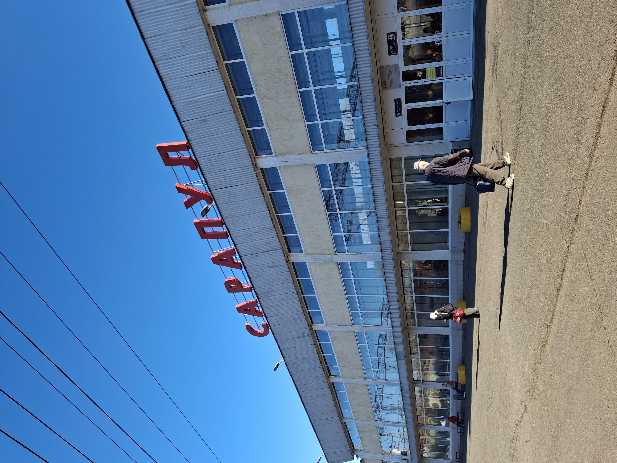 The image shows a tilted view of a building in Sarapul, Russia, identified by the large red Cyrillic letters spelling "САРАПУЛ" attached to the building's overhang. The building has multiple windows and doors, reflecting the bright blue sky. An elderly man in a dark jacket, dark pants, white shoes, and a white hat is walking on the pavement in front of the building, carrying a dark bag. The pavement is asphalt. There are yellow cylindrical objects near the building's entrance. Power lines stretch across the sky in the background.