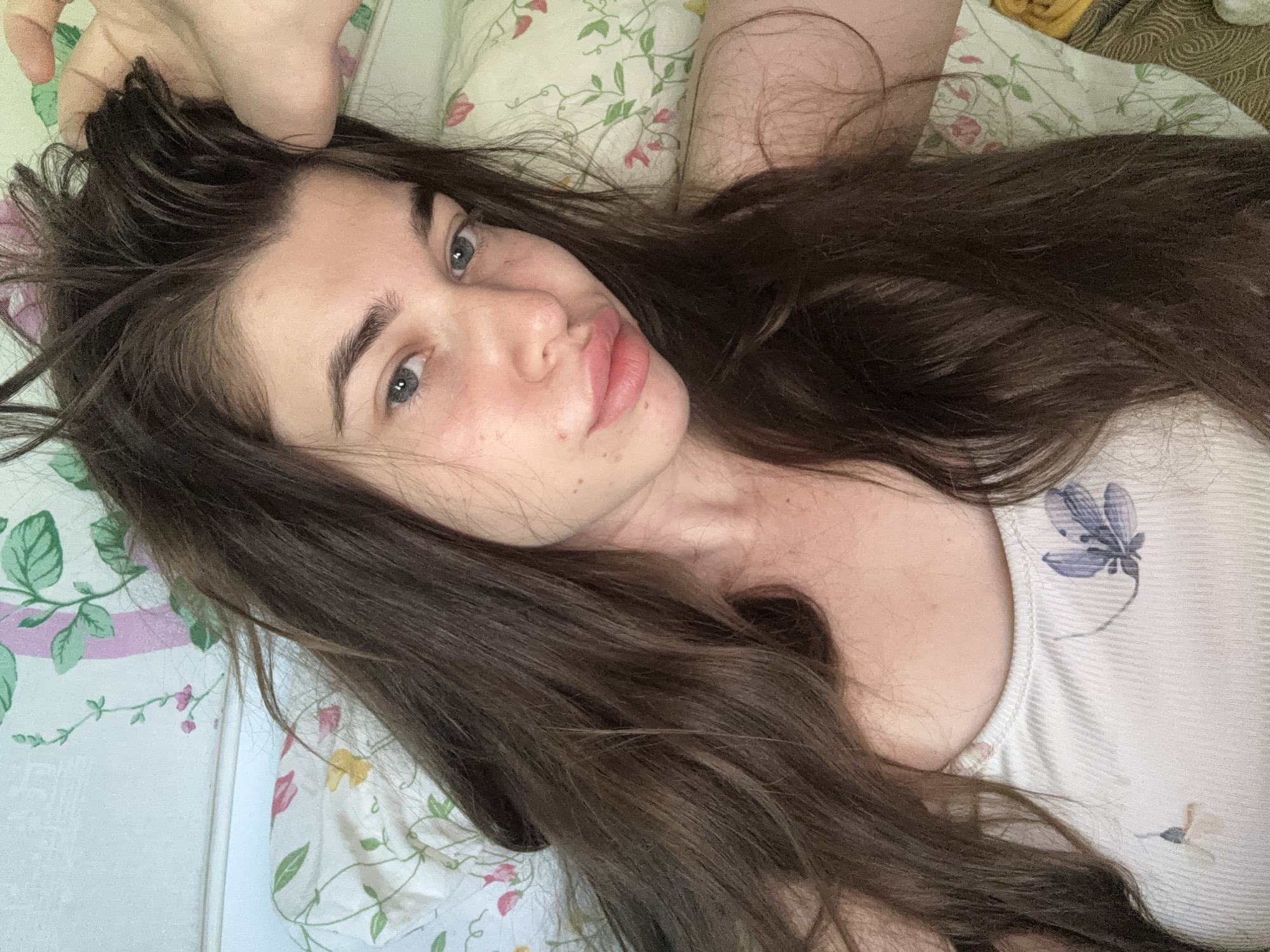 The image shows a close-up, overhead shot of a young woman lying in bed, looking at the camera. She has long, dark brown hair that is messy and covering part of her face. Her eyes are blue, and she has full lips. She is wearing a white tank top with a floral pattern. The background appears to be a wall with a floral wallpaper and a pillow with a similar pattern. The lighting suggests it is daytime. The scene is indoors, likely a bedroom. There is no visible indication of the location being in the city of Мещёры, Russia.