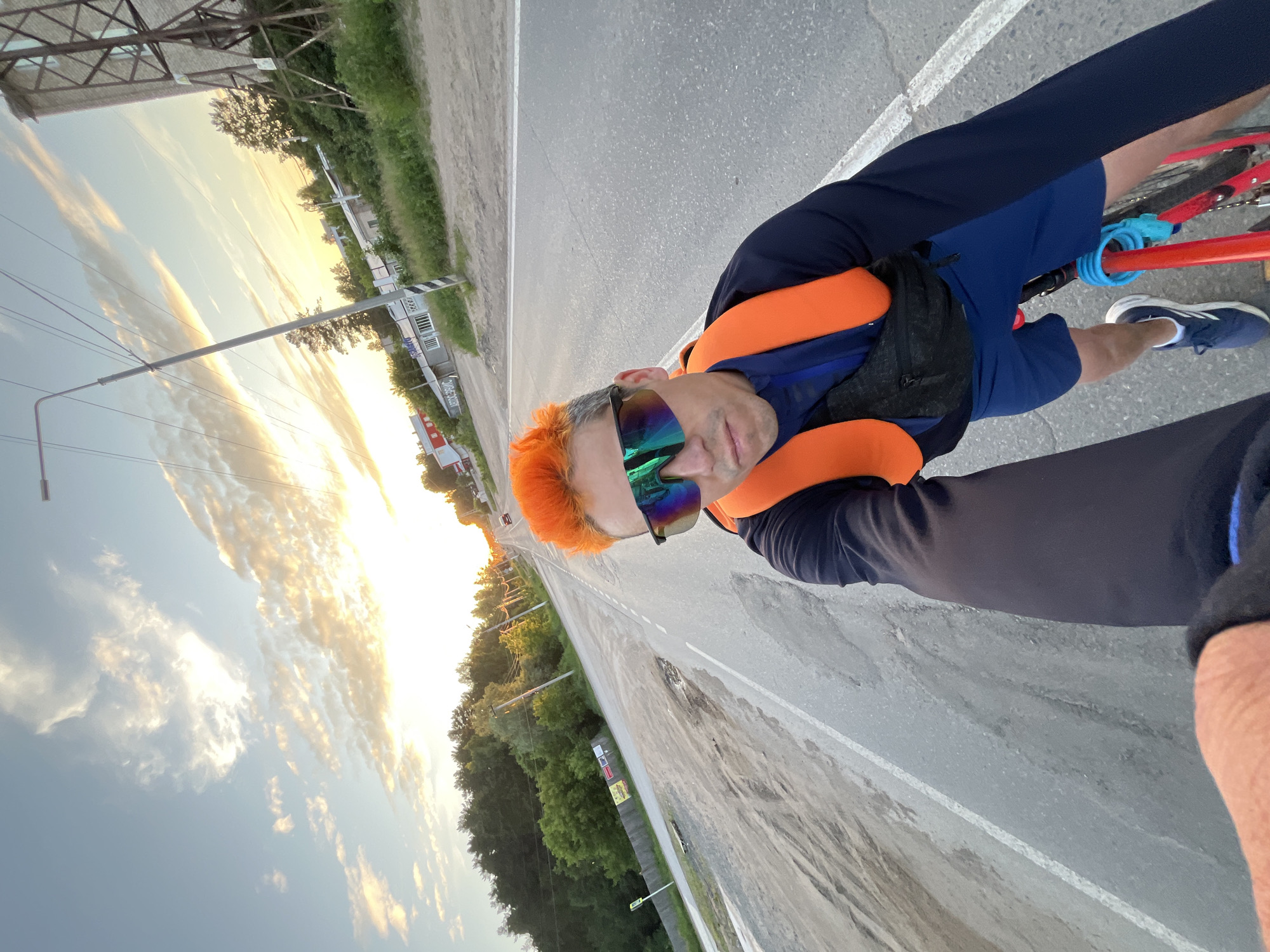 This image depicts a male individual on a bicycle on a road in Chaikovsky, Russia, during what appears to be either sunrise or sunset. The man has distinct bright orange, spiky hair and wears large, reflective, multi-colored sunglasses. He is dressed in a dark long-sleeved shirt, bright orange padded shoulder straps (suggesting a backpack or vest), a black fanny pack worn across his chest, dark blue shorts, and dark blue athletic shoes with white soles. He is holding the camera with his extended left arm, taking a selfie. Parts of a red bicycle frame with a turquoise-blue cable lock are visible beneath him.
The asphalt road features a white solid line on the right shoulder and extends into the distance, where a light-colored vehicle is visible. The road surface shows signs of wear and tear. On the left side of the road, there is a dirt shoulder with visible tire tracks, leading to a wooden fence, several partially visible signs or billboards, and dense green trees and bushes. Beyond the vegetation, commercial buildings are present, including one with "АВТОСЕРВИС" (AUTOSERVICE) and another with "МАГАЗИН" (SHOP) in Cyrillic lettering. A street lamp and overhead power lines run parallel to the road on this side. On the right side, there is a similar dirt shoulder, green vegetation, a tall metal lattice structure (possibly a water tower), and further buildings in the background. The sky displays a mix of clear blue and scattered clouds, illuminated by the low sun with warm, golden and orange hues.