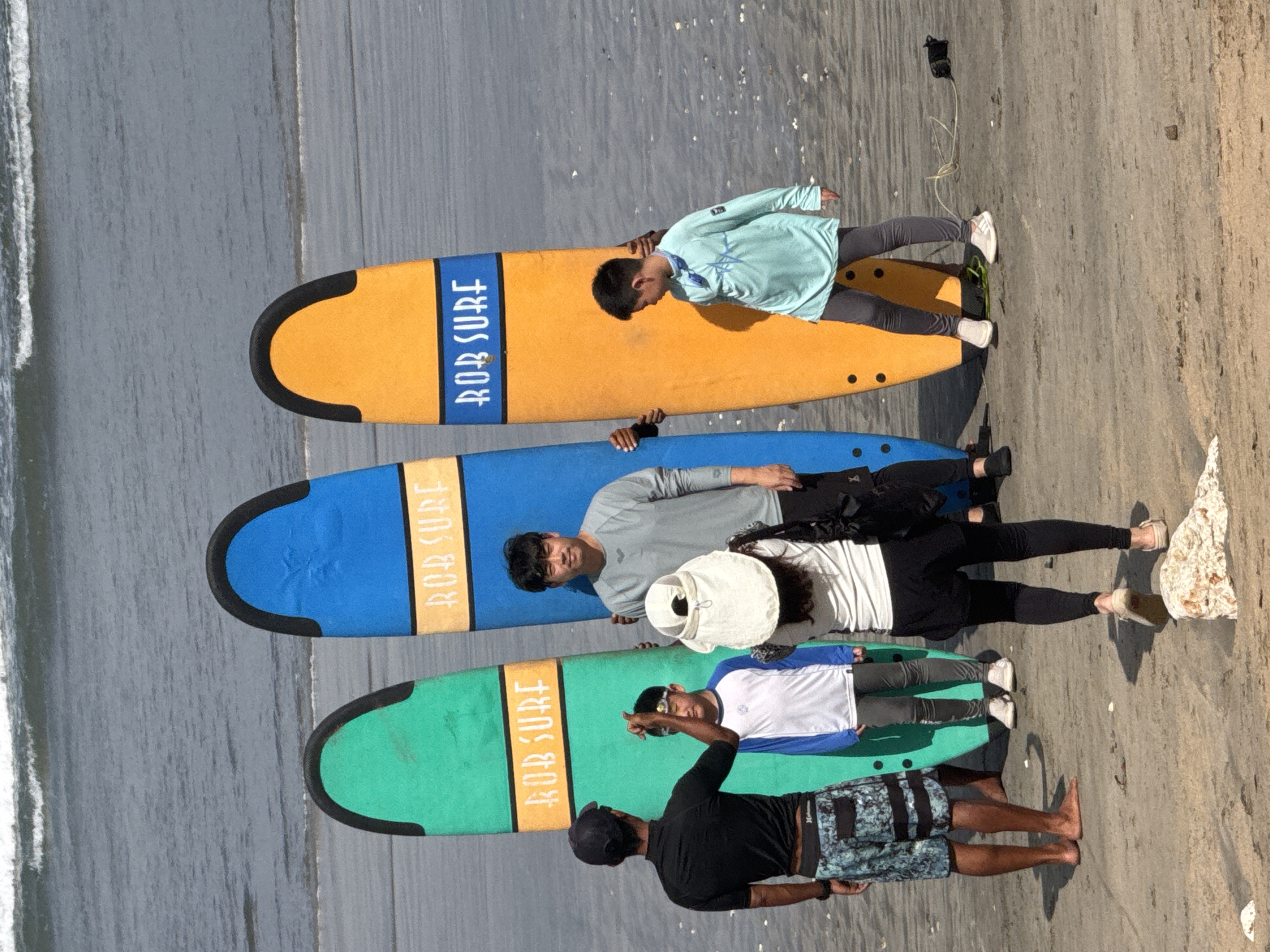 The image depicts a beach scene in Kuta, Indonesia, where five individuals are gathered with three surfboards. In the foreground, a woman wearing a white hat and a black outfit is facing away from the camera, holding a smartphone and appearing to take a photograph. To her left, a man in a black t-shirt and patterned shorts stands facing the group, with his right arm raised. Beside him, a young boy in a blue and white long-sleeved shirt and grey pants looks towards the ground. Behind the boy, a blue surfboard stands upright, emblazoned with "ROB SURF". To the right of the blue surfboard, a man in a grey long-sleeved shirt stands holding the surfboard. To the right of this man, an orange surfboard is also standing upright, also bearing the "ROB SURF" inscription. Another young boy in a light blue long-sleeved shirt and grey pants stands in front of the orange surfboard, looking down. In the background, the ocean with moderate waves is visible, and the sandy beach stretches towards the water. The overall atmosphere suggests a casual outing or surf lesson.