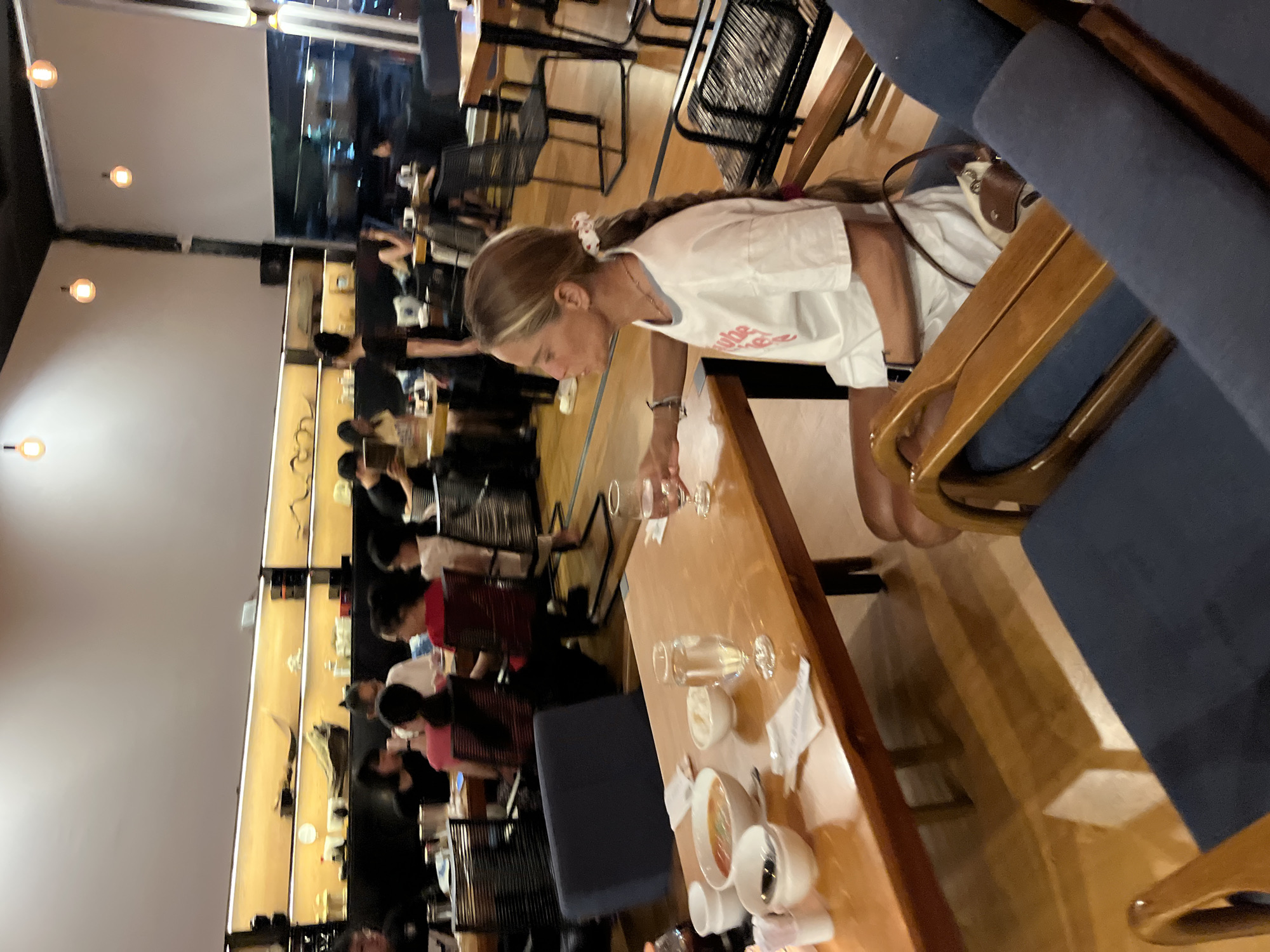 The image depicts an indoor dining scene, likely a restaurant, with several tables and patrons. In the foreground, a woman with long braided hair is seated at a table, wearing a white t-shirt. The table has several bowls of food, a glass of water, and chopsticks. The background shows other diners, some of whom appear to be young people. The restaurant has wooden furniture and modern lighting fixtures. The location is identified as Phu Thuy Ward, Vietnam, suggesting a setting within that area. The overall ambiance appears to be casual and social.