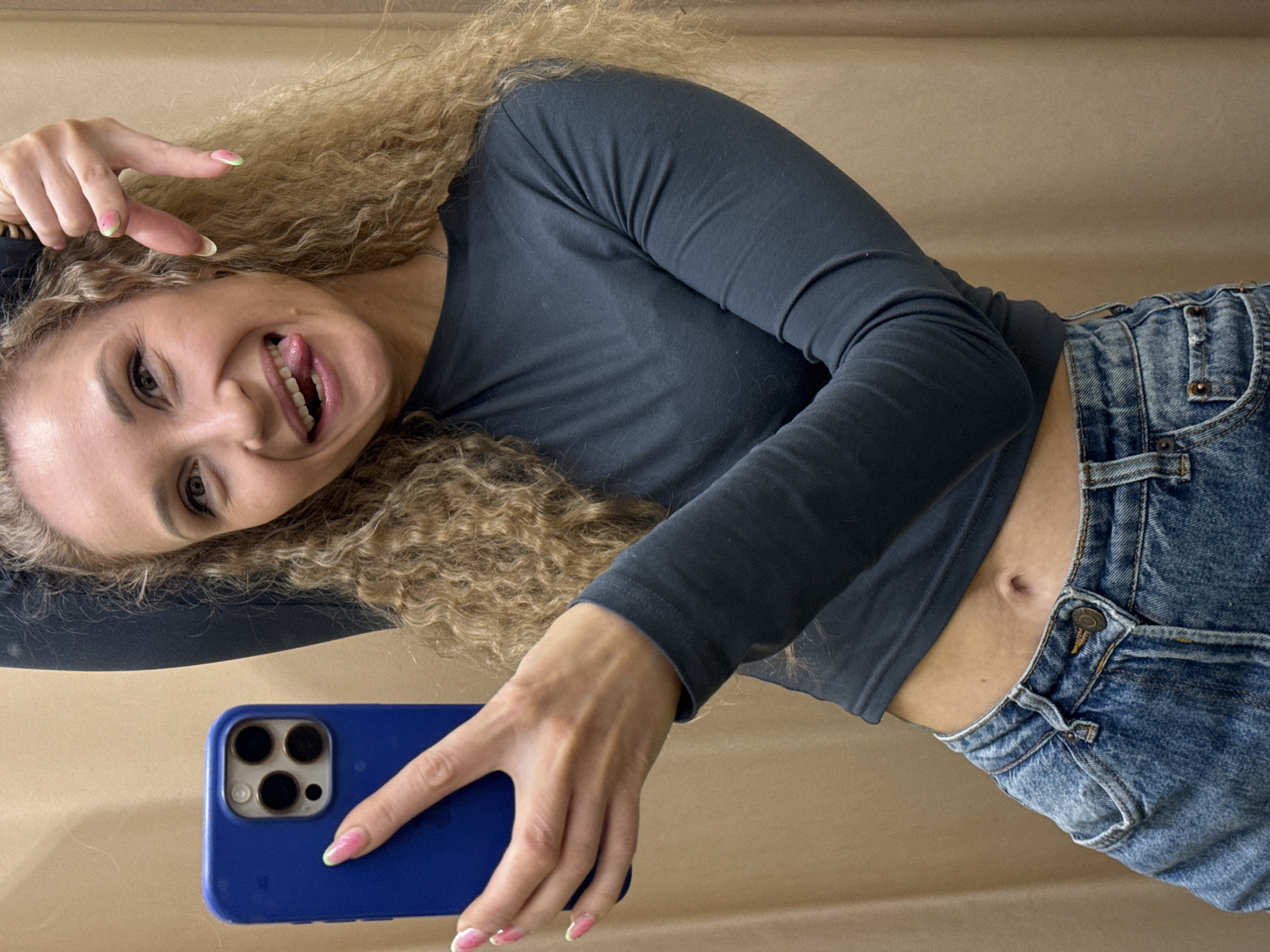 A young woman with curly blonde hair is taking a selfie with a blue smartphone. She is wearing a dark gray long-sleeved shirt and blue jeans. She is sticking her tongue out and making a peace sign with her left hand near her face. Her nails are painted with a pink and green watermelon design. The background is a light brown, plain surface. There is no indication in the image that it was taken in the city of Perm, Russia.