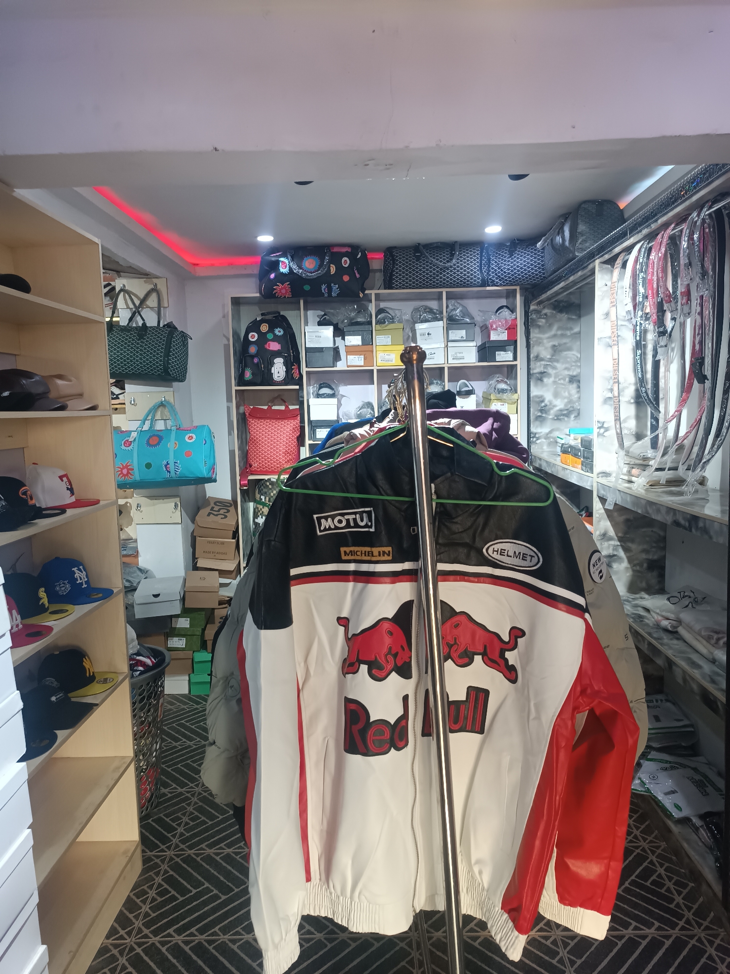 The image depicts the interior of a retail space, possibly a clothing store, in Bukuru, Nigeria. The foreground showcases a rack of clothing, dominated by a white jacket with red accents and a "Red Bull" logo, along with racing-themed branding such as "MOTU" and "MICHELIN" patches, and "HELMET." The rack is located on a patterned floor.
To the left, a shelf unit contains a variety of items, including hats and boxed goods. Behind the jacket rack, bags, backpacks, and boxes are stacked on shelves. Along the right side of the frame, there's a section with shelving and hanging items.
The overall lighting is a mix of natural and artificial light, with some red accent lighting near the ceiling. The space is somewhat crowded, suggesting a small business operation.