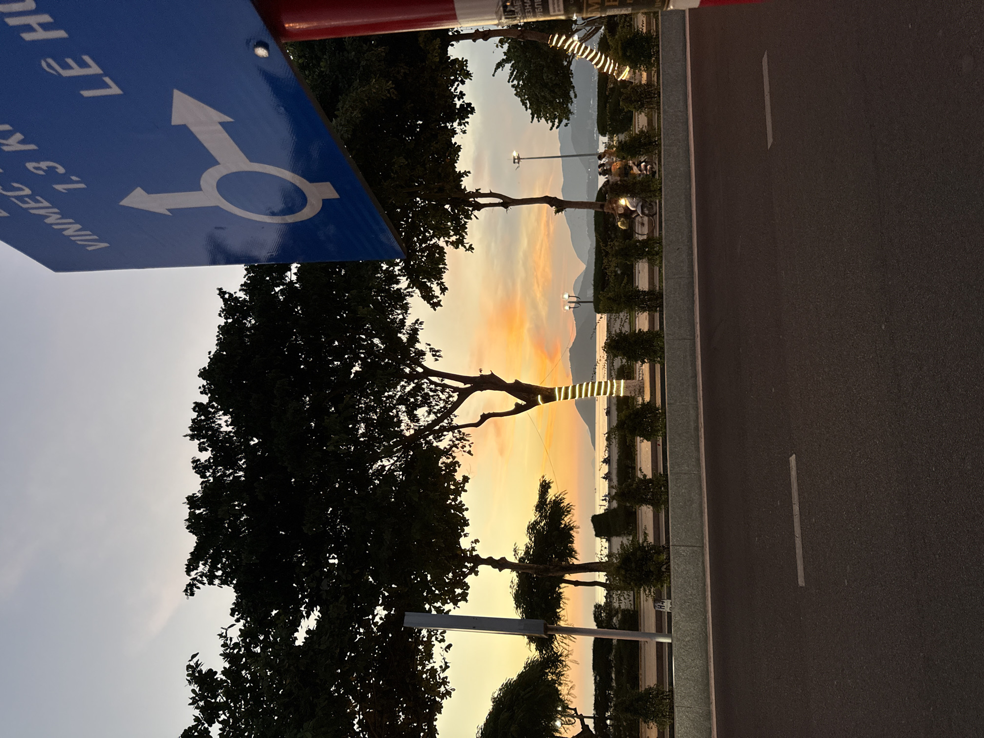 The image captures a street scene in Nha Trang, Vietnam, during twilight. A blue traffic sign with white arrows indicating a roundabout and text reading "VINMEC 1,3 K" and "LÊ HO" is prominently displayed on the right. Beyond the road, trees with dark foliage line a waterfront promenade decorated with string lights. In the background, the calm sea is visible, with hills and mountains forming the horizon. The sky displays the warm hues of sunset. A few figures are seen on a motorcycle on the promenade, and a lamppost stands tall amongst the trees. The foreground features an asphalt road with faint white markings.