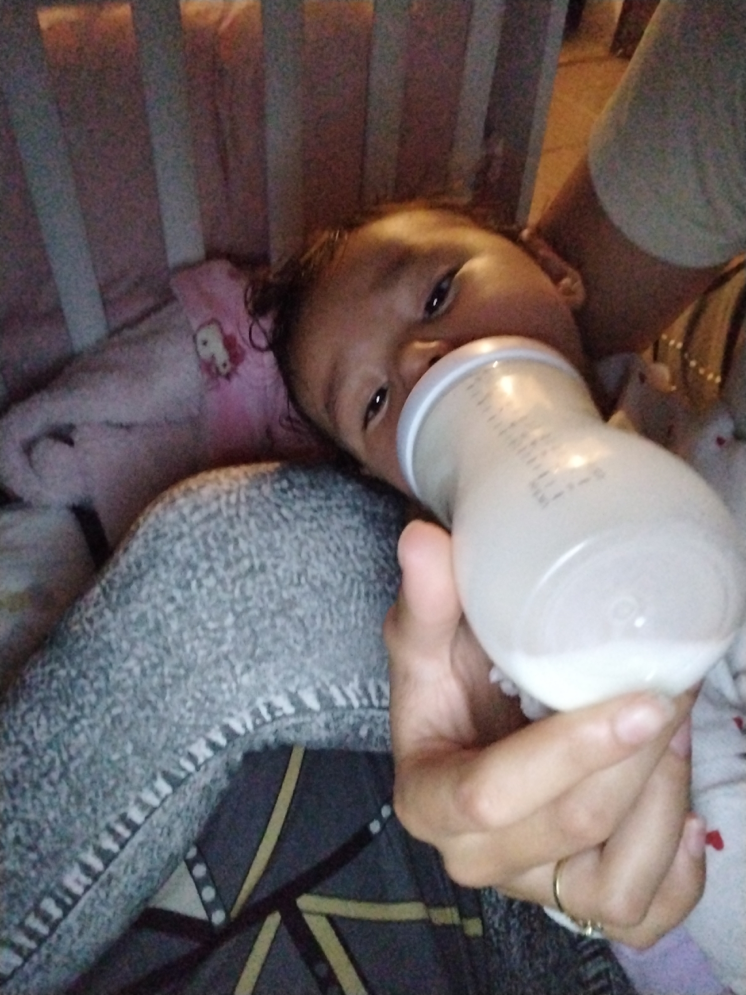 A baby is lying on its back in a crib, being bottle-fed milk by a person. The baby is holding the bottle in their hands. The crib has white bars and a pink blanket with a Hello Kitty design. The person holding the bottle is wearing a light gray t-shirt. The bottom sheet of the crib is gray and black with a geometric pattern. The location is Unknown, Unknown.