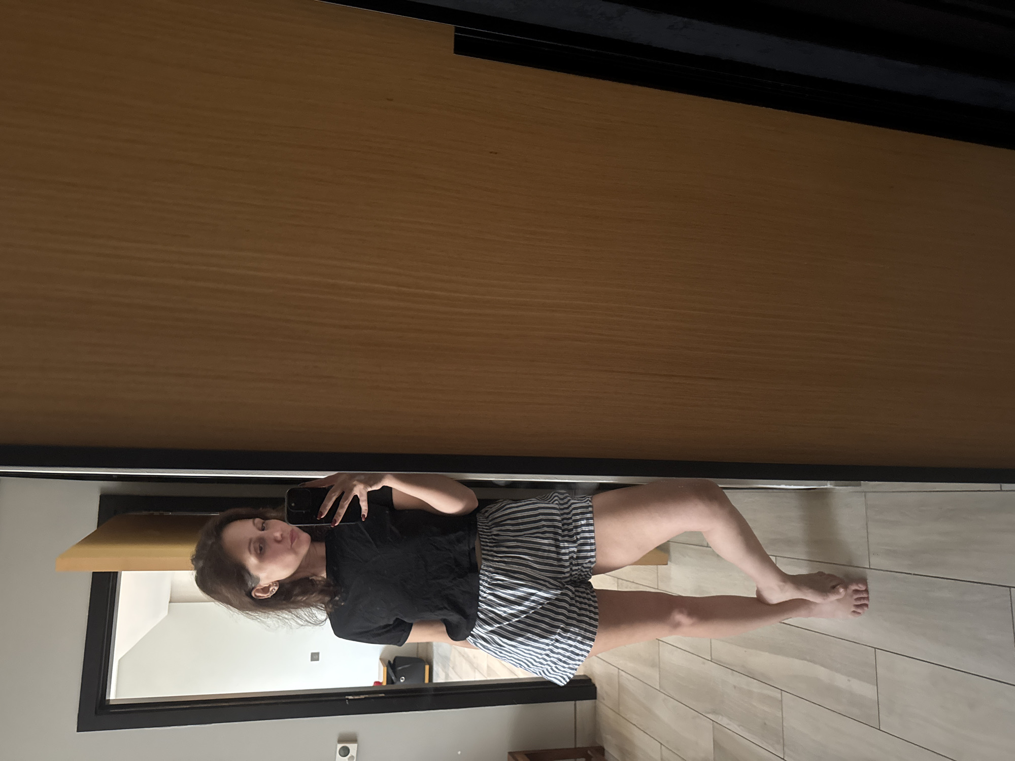 A woman is taking a selfie in a mirror. She is wearing a black t-shirt and striped shorts. The background shows a room with tiled floors and light-colored walls. The location is a room, possibly a residence, but there is no evidence to suggest it is located in Dubai or the United Arab Emirates.