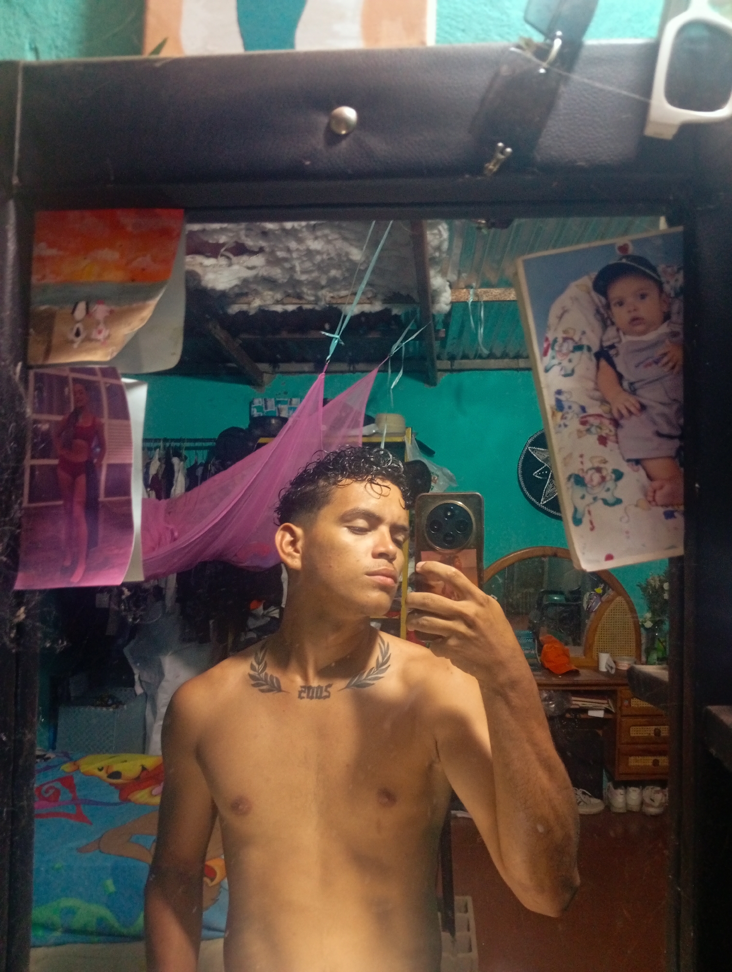 A shirtless young man is taking a selfie in a mirror. The mirror has a dark frame and is adorned with various items including a photo of a baby, a picture of a woman in a bathing suit, and what appears to be a drawing or postcard with a sunset scene. The room reflected in the mirror seems cluttered, with clothes hanging, a pink mosquito net, and a cartoon-themed blanket. Furniture is visible, including a wooden desk with objects on top and a chair nearby. The wall is painted turquoise. The tattoos visible on the man's chest indicate the number 2005. The city of Altagracia, Venezuela cannot be confirmed by the image provided.