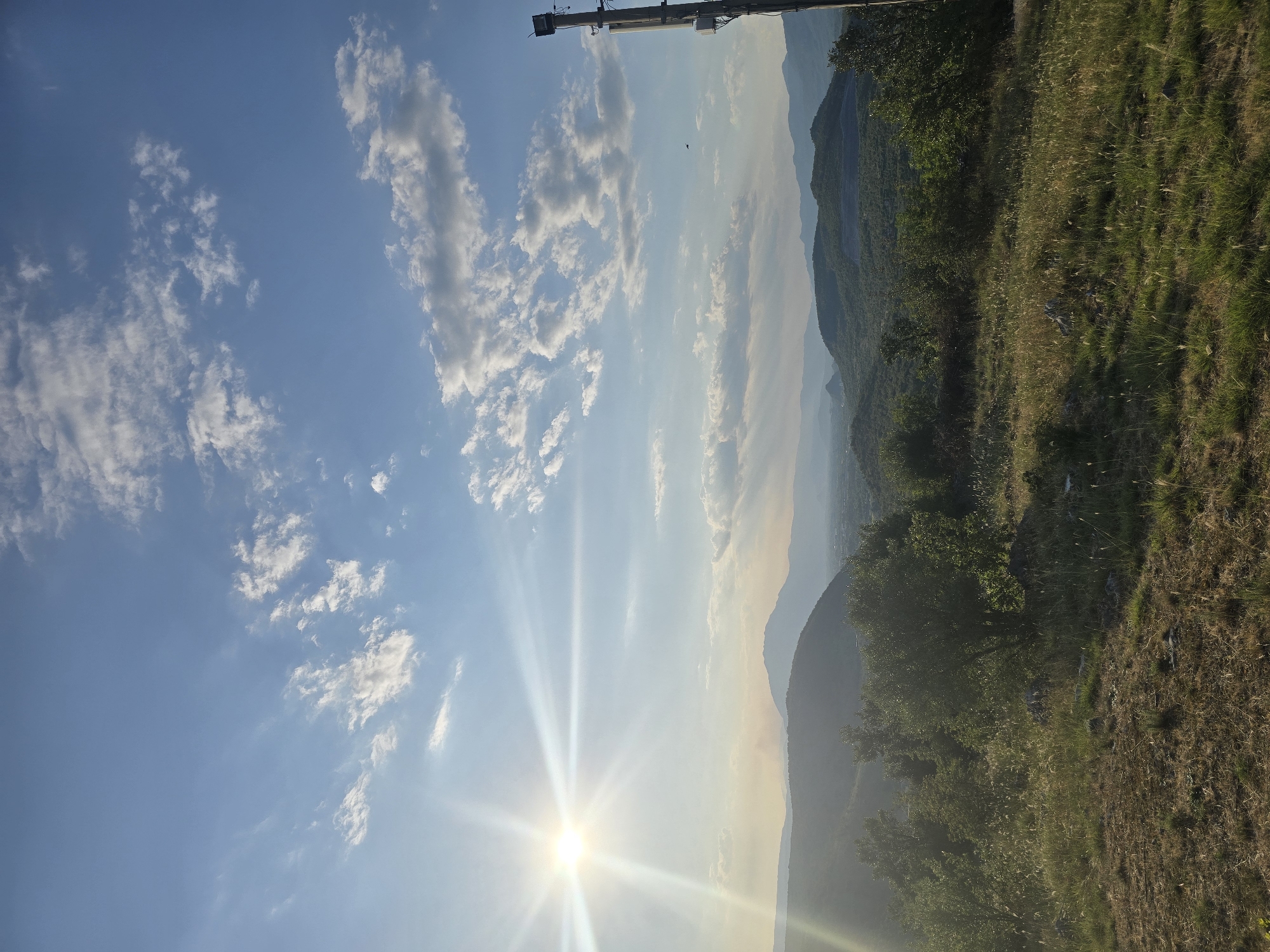 A vertical, eye-level, wide-angle shot captures a sun-drenched, hilly landscape under a bright blue sky with scattered clouds. The sun, positioned in the upper left quadrant, emits strong rays and lens flare, illuminating the scene. Rolling hills covered in dense green foliage and trees dominate the midground and background, fading into a hazy horizon. In the foreground, dry grass and patches of darker earth suggest an arid or late-summer environment. A solitary, tall utility pole with a light fixture and equipment is visible on the far right edge of the frame. The overall atmosphere is one of serene natural beauty, likely captured during late afternoon or early morning due to the quality of light. The location is identified as Unknown, Unknown.