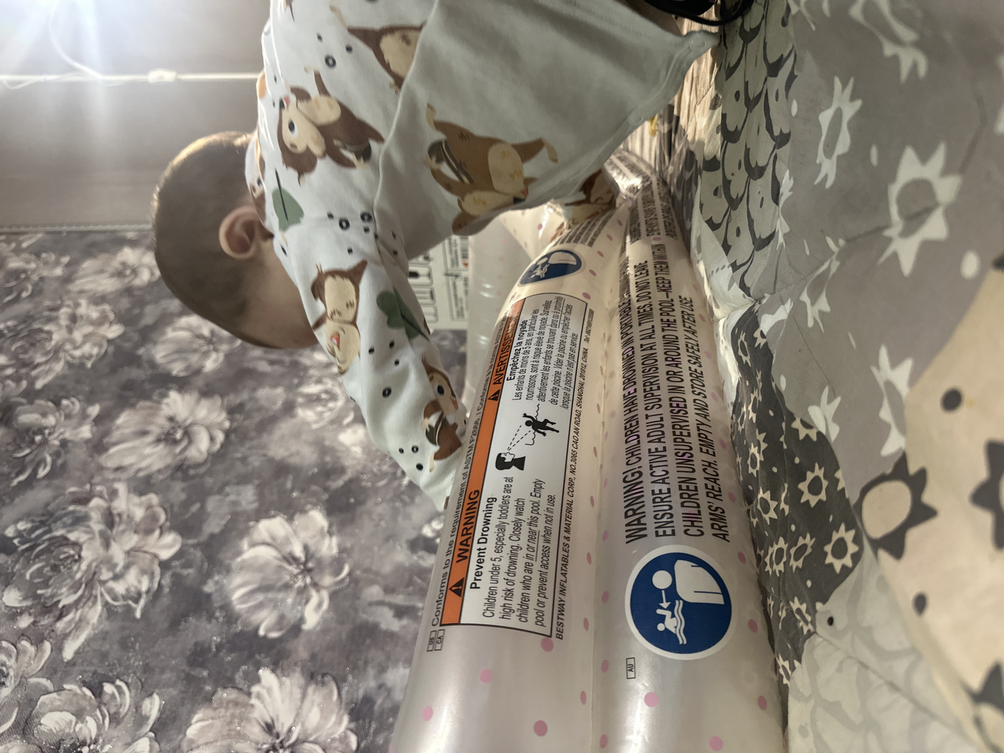 A close-up shot shows a young child in a long-sleeved outfit with a monkey pattern, peering at two inflatable pool floats. The floats are pink with white labels displaying drowning prevention warnings in both English and French. The label includes the name BESTWAY INFLATABLES & MATERIAL CORP., with an address in Shanghai, China. The background features a gray floral pattern on what appears to be bedding or a surface covering.