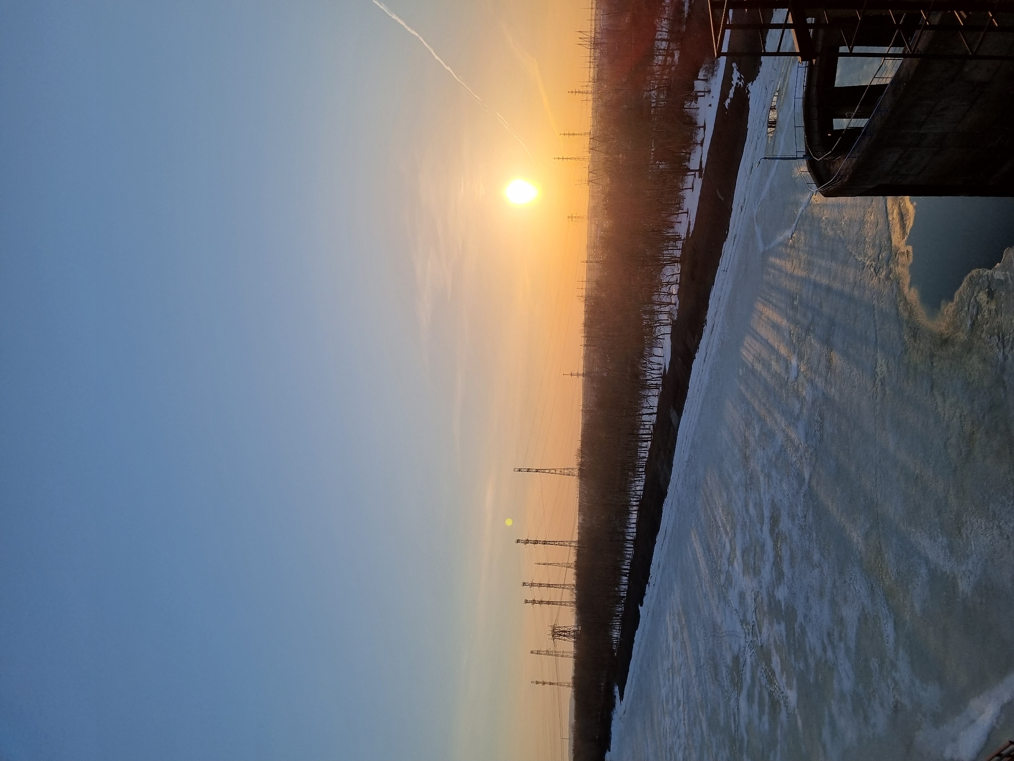 The image shows a sunset over the Kama River in Chaikovsky, Russia. The river is mostly frozen with patches of open water. Power lines are visible along the riverbank. A structure, possibly part of a power plant or industrial facility, is visible on the right. The sky transitions from blue to orange near the horizon where the sun is setting. A contrail is visible in the upper left of the sky.