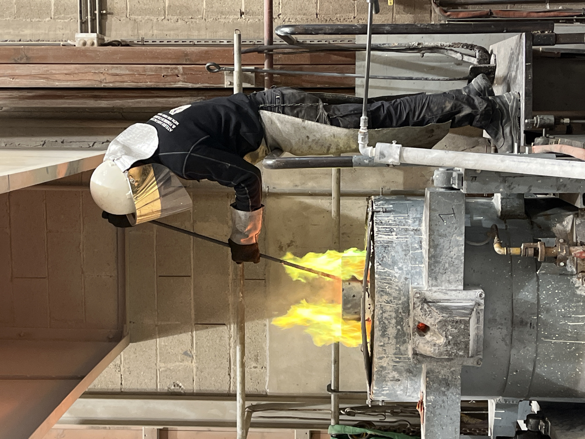 A person wearing protective gear, including a helmet and gloves, is positioned horizontally in front of a furnace or industrial oven emitting bright yellow and green flames. The person is holding a long metal rod, possibly interacting with the contents of the furnace. The scene appears to be inside an industrial facility with exposed piping and brick or concrete walls. The person is wearing dark clothing and has a protective apron or covering over their legs. The jacket has the words "ATELIER FABRICATION" and "METAUX & FONDERIE D" written on it. The location is Saint-Rémy-lès-Chevreuse, France.