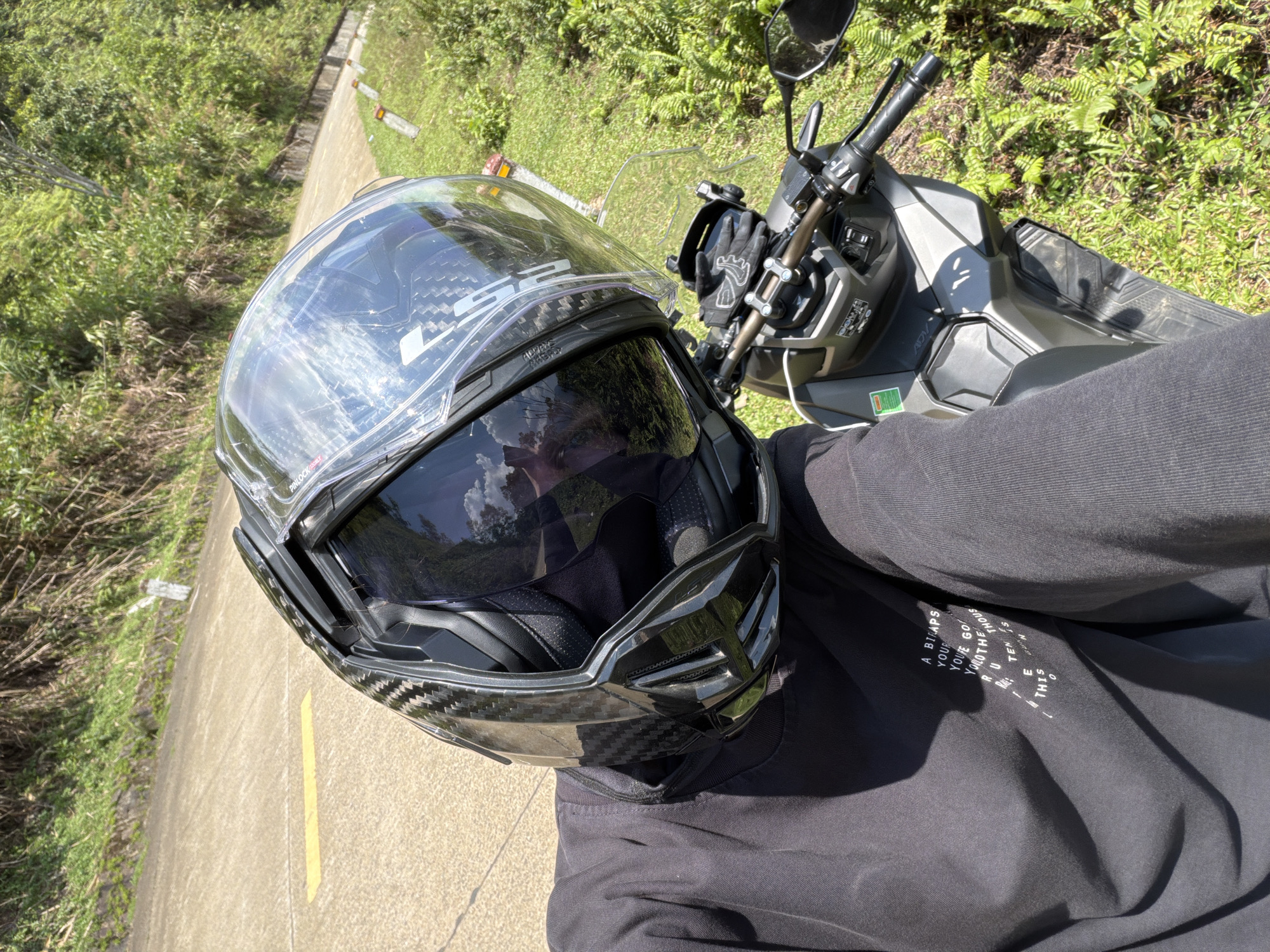 The image shows an individual sitting on a motorcycle, wearing a full-face motorcycle helmet with a dark visor. The helmet is black and appears to have a carbon fiber pattern. The individual is wearing a long-sleeved black shirt with white text on the lower left side, partially obscured by folds. The motorcycle has a gray or silver color scheme. The background consists of greenery and a paved road with a yellow line. The location is likely a rural or semi-rural area in the city of Huế, Vietnam.